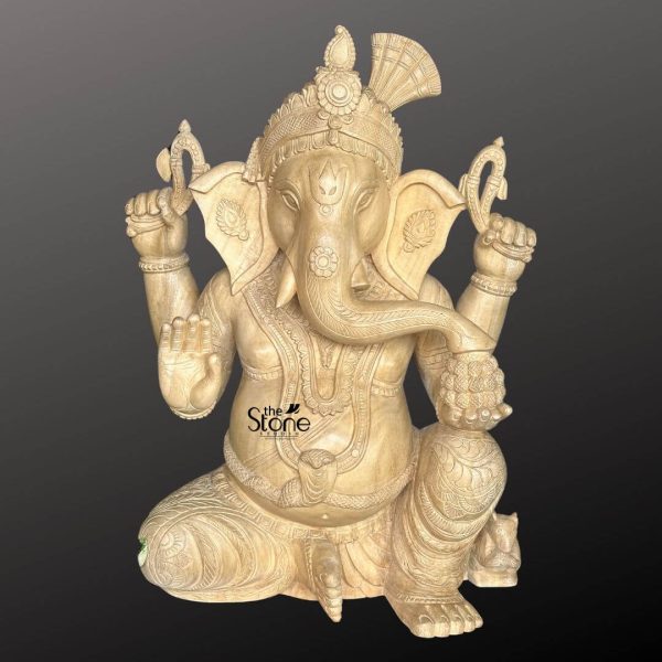 A beautifully carved 2-foot wooden Ganesha statue is presented from a frontal slightly elevated perspective against a plain dark background, highlighting its intricate details for online viewing. The statue is crafted from light-colored wood, showcasing its natural grain and warm earthy tones. Lord Ganesha is depicted in a traditional seated posture with one knee raised exuding an aura of wisdom, prosperity and benevolence. Ganesha is shown with four arms his upper hands hold traditional attributes, possibly a noose and an axe. His lower right hand is raised in a gesture of blessing offering protection while his lower left hand rests on his lap possibly holding a modak. His elephant head is intricately detailed with large fan-like ears expressive eyes and his trunk curving gracefully towards his left. He wears an elaborate crown adorned with a fan-shaped element at the back along with intricate necklaces armlets, and a sacred thread across his plump belly. His garments are finely sculpted with delicate folds and patterns enhancing the traditional aesthetic. A small mouse, Ganesha's vehicle is subtly carved near his foot, though partially obscured. The natural wood finish adds to its rustic and handcrafted charm. This detailed image vividly conveys the spiritual majesty and artistic craftsmanship of the wooden Ganesha statue making it highly relevant to an article discussing online availability of such idols.