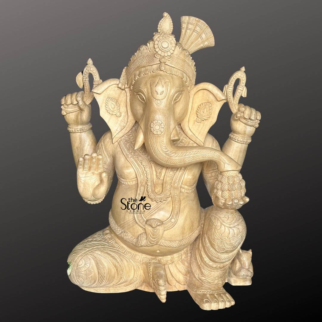 Wooden Ganesha Statue Online 2ft 1 A beautifully carved 2-foot wooden Ganesha statue is presented from a frontal slightly elevated perspective against a plain dark background, highlighting its intricate details for online viewing. The statue is crafted from light-colored wood, showcasing its natural grain and warm earthy tones. Lord Ganesha is depicted in a traditional seated posture with one knee raised exuding an aura of wisdom, prosperity and benevolence. Ganesha is shown with four arms his upper hands hold traditional attributes, possibly a noose and an axe. His lower right hand is raised in a gesture of blessing offering protection while his lower left hand rests on his lap possibly holding a modak. His elephant head is intricately detailed with large fan-like ears expressive eyes and his trunk curving gracefully towards his left. He wears an elaborate crown adorned with a fan-shaped element at the back along with intricate necklaces armlets, and a sacred thread across his plump belly. His garments are finely sculpted with delicate folds and patterns enhancing the traditional aesthetic. A small mouse, Ganesha's vehicle is subtly carved near his foot, though partially obscured. The natural wood finish adds to its rustic and handcrafted charm. This detailed image vividly conveys the spiritual majesty and artistic craftsmanship of the wooden Ganesha statue making it highly relevant to an article discussing online availability of such idols.