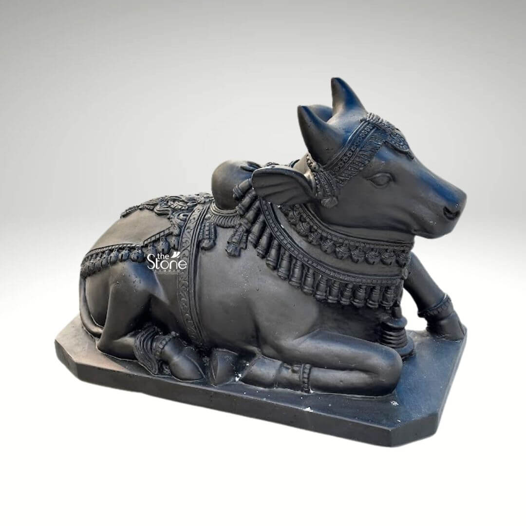 Black Nandi Bull Statue for Home 2.5ft 2 A majestic 2.5-foot black stone Lord Shiva Nandi statue is prominently featured against a clean neutral background emphasizing its intricate details and serene presence. This exquisite sculpture depicts Nandi the revered vahana of Lord Shiva in a classic reclining posture symbolizing unwavering patience devotion, and strength making it ideal for an article discussing such sacred statues. The bull is shown lying down with its legs gracefully folded beneath its body its head turned slightly to the right looking forward with a calm and benevolent expression. The craftsmanship of the statue is exceptional showcasing the smooth deep black stone that beautifully accentuates Nandi's powerful yet serene form. The bull is richly adorned with meticulously carved traditional Indian jewelry and ceremonial embellishments. These include an elaborate crown or headpiece on its forehead intricately designed necklaces with dangling ornaments around its neck and decorative blankets or saddle covers carved onto its back all integrated seamlessly into the stone. The details of the ornaments such as beads bells and patterns are finely rendered enhancing the statue's divine appearance and cultural authenticity. Nandi's facial features are finely sculpted with a tranquil and benevolent expression characterized by gentle eyes a strong snout and well-formed horns that rise gracefully from its head. The texture of the black stone with its inherent depth and subtle sheen gives the statue a timeless and dignified quality. The bull rests on a sturdy rectangular black stone base which provides stability and a formal presentation. This 2.5-foot blackstone Nandi statue is not merely a decorative item but a powerful spiritual artifact ideal for a temple home temple entrance, or garden inspiring devotion and bringing an auspicious presence to the living space.