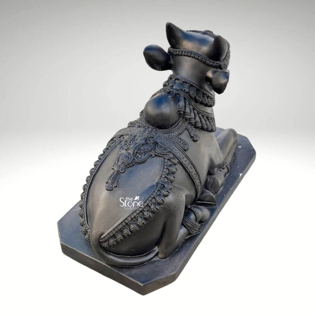 Black Nandi Bull Statue for Home 2.5ft 4 A majestic 2.5-foot black stone Nandi idol specifically crafted for worship is prominently featured from a slightly elevated rear-side perspective against a clean neutral background emphasizing its intricate details and sacred presence. This exquisite sculpture depicts Nandi the revered vahana of Lord Shiva in a classic reclining posture symbolizing unwavering patience devotion and strength making it ideal for an article discussing sacred Nandi idols for worship. The bull is shown lying down with its legs gracefully folded beneath its body its head turned slightly to the right looking forward with a calm and benevolent expression even from this angle. The craftsmanship of the idol is exceptional showcasing the smooth deep black stone that beautifully accentuates Nandi's powerful yet serene form. The bull is richly adorned with meticulously carved traditional Indian jewelry and ceremonial embellishments. These include an elaborate crown or headpiece on its forehead intricately designed necklaces with dangling ornaments around its neck and decorative blankets or saddle covers carved onto its back all integrated seamlessly into the stone. The details of the ornaments such as beads bells and patterns are finely rendered enhancing the idol's divine appearance and cultural authenticity. From this view the intricate patterns on Nandi's back and sides are particularly prominent outlined by decorative borders. Nandi's facial features though partially visible still convey a sense of tranquility. His well-formed horns rise gracefully from his head. The texture of the black stone with its inherent depth and subtle sheen gives the idol a timeless and dignified quality suitable for veneration. The bull rests on a sturdy rectangular black stone base which provides stability and a formal presentation consistent with an object of worship. This 2.5-foot blackstone Nandi idol is not merely a decorative item but a powerful spiritual artifact ideal for a temple home temple or sacred space inspiring deep devotion and bringing an auspicious presence for religious ceremonies.