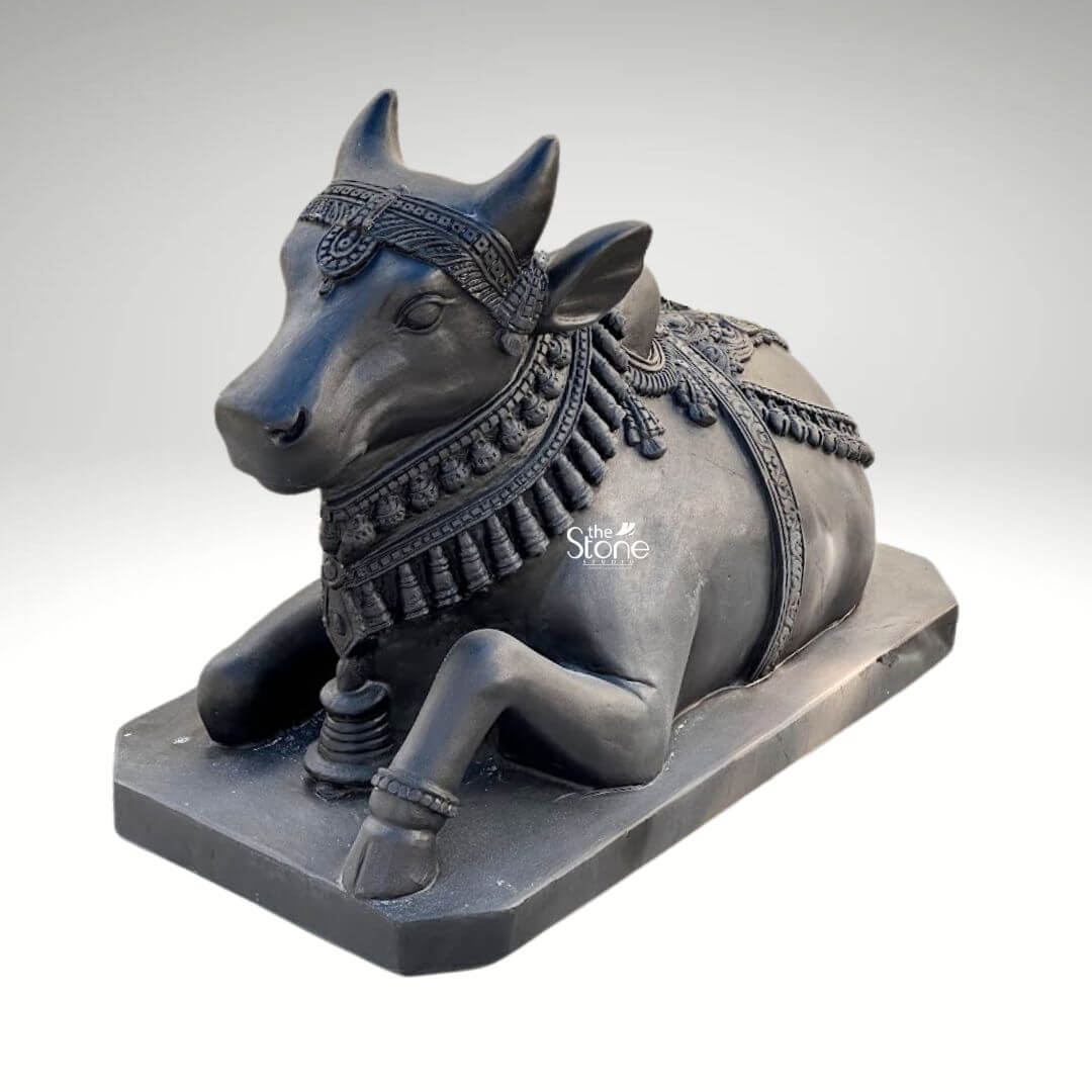 Black Nandi Bull Statue for Home 2.5ft 5 A magnificent 2.5-foot handcrafted black stone Nandi Bull statue is prominently featured against a clean neutral background emphasizing its intricate details and serene presence. This exquisite sculpture depicts Nandi the revered vahana of Lord Shiva in a classic reclining posture symbolizing unwavering patience devotion and strength making it ideal for an article discussing such handcrafted statues. The bull is shown lying down with its legs gracefully folded beneath its body its head turned slightly to the right looking forward with a calm and benevolent expression. The craftsmanship of the handcrafted statue is exceptional showcasing the smooth deep black stone that beautifully accentuates Nandi's powerful yet serene form. The bull is richly adorned with meticulously carved traditional Indian jewelry and ceremonial embellishments. These include an elaborate crown or headpiece on its forehead intricately designed necklaces with dangling ornaments around its neck and decorative blankets or saddle covers carved onto its back all integrated seamlessly into the stone. The details of the ornaments such as beads bells and patterns are finely rendered enhancing the statue's divine appearance and cultural authenticity highlighting the skill of the artisan. Nandi's facial features are finely sculpted with a tranquil and benevolent expression characterized by gentle eyes a strong snout and well-formed horns that rise gracefully from its head. The texture of the black stone with its inherent depth and subtle sheen gives the statue a timeless and dignified quality. The bull rests on a sturdy rectangular black stone base which provides stability and a formal presentation. This 2.5-foot handcrafted blackstone Nandi Bull statue is not merely a decorative item but a powerful spiritual artifact ideal for a temple home temple entrance, or garden inspiring devotion and bringing an auspicious presence to any space.