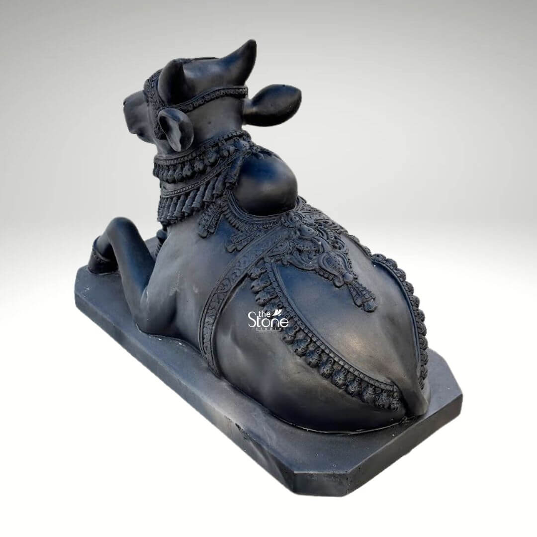 Black Nandi Bull Statue for Home 2.5ft 6 A majestic 2.5-foot black stone Nandi statue specifically crafted for an outdoor temple setting is prominently featured from a slightly elevated rear-side perspective against a clean neutral background emphasizing its intricate details and sacred presence. This exquisite sculpture depicts Nandi the revered vahana of Lord Shiva in a classic reclining posture symbolizing unwavering patience devotion and strength making it ideal for an article discussing such sacred statues for outdoor temples. The bull is shown lying down with its legs gracefully folded beneath its body its head turned slightly to the right looking forward with a calm and benevolent expression even from this angle. The craftsmanship of the statue is exceptional showcasing the smooth deep black stone that beautifully accentuates Nandi's powerful yet serene form. The bull is richly adorned with meticulously carved traditional Indian jewelry and ceremonial embellishments. These include an elaborate crown or headpiece on its forehead intricately designed necklaces with dangling ornaments around its neck, and decorative blankets or saddle covers carved onto its back all integrated seamlessly into the stone. The details of the ornaments, such as beads bells and patterns, are finely rendered enhancing the statue's divine appearance and cultural authenticity. From this view the intricate patterns on Nandi's back and sides outlined by decorative borders are particularly prominent emphasizing the elaborate craftsmanship. Nandi's facial features though partially visible still convey a sense of tranquility. His well-formed horns rise gracefully from his head. The texture of the black stone with its inherent depth and subtle sheen gives the statue a timeless and dignified quality suitable for veneration in an outdoor temple where it would withstand the elements. The bull rests on a sturdy rectangular black stone base which provides stability and a formal presentation consistent with an object of worship. This 2.5-foot blackstone Nandi statue is not merely a decorative item but a powerful spiritual artifact, inspiring deep devotion and bringing an auspicious presence to any outdoor temple or sacred space.