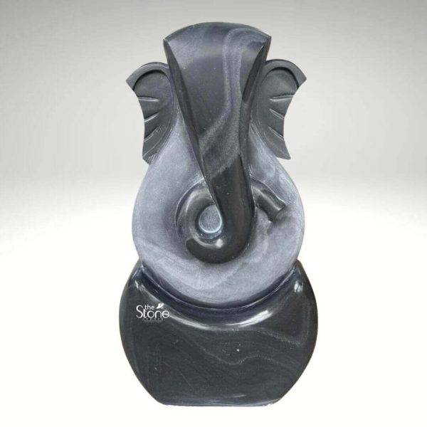 A striking three-foot abstract Ganesha statue meticulously crafted from black stone is presented against a neutral background emphasizing its modern artistry and symbolic form. This contemporary interpretation of Lord Ganesha, ideal for an article discussing abstract Ganesha sculptures features a minimalist yet recognizable representation of the beloved deity. The statue predominantly uses sweeping smooth curves and simplified forms to suggest Ganesha's iconic elephant head trunk and ears rather than a literal depiction. The sculpture utilizes two contrasting tones of black stone creating a dynamic visual effect. The outer darker black stone forms the core outline of Ganesha's head and a rounded base. Within this a lighter subtly textured gray-black stone highlights the central features particularly defining the elongated trunk that elegantly curls to the left and the broad fan-like ears. The interplay of these two dark tones adds depth and sophistication to the abstract design allowing light to catch the varying surfaces and reveal subtle striations in the stone. The overall form is vertical and streamlined with the top suggesting Ganesha's crown and the bottom forming a stable rounded base. Despite its abstract nature the essence of Ganesha — wisdom prosperity and the remover of obstacles — is clearly conveyed through the familiar contours. This three-foot blackstone abstract Ganesha statue is a contemporary work of art that seamlessly blends spiritual symbolism with modern design making it a unique and powerful addition to any modern home office, or art collection as suggested by its context.