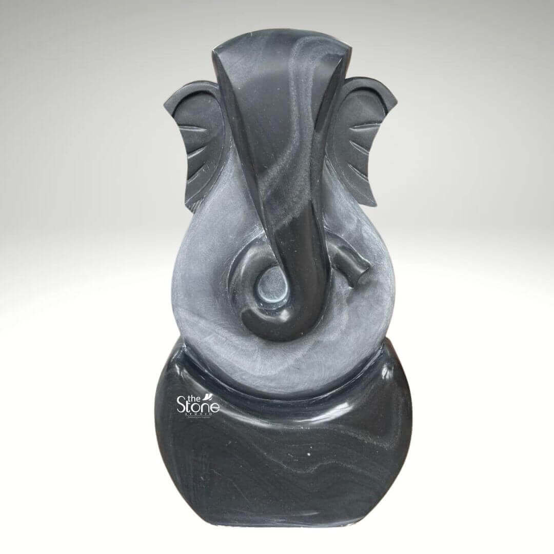 3 Feet Abstract Ganesha Statue 1 A striking three-foot abstract Ganesha statue meticulously crafted from black stone is presented against a neutral background emphasizing its modern artistry and symbolic form. This contemporary interpretation of Lord Ganesha, ideal for an article discussing abstract Ganesha sculptures features a minimalist yet recognizable representation of the beloved deity. The statue predominantly uses sweeping smooth curves and simplified forms to suggest Ganesha's iconic elephant head trunk and ears rather than a literal depiction. The sculpture utilizes two contrasting tones of black stone creating a dynamic visual effect. The outer darker black stone forms the core outline of Ganesha's head and a rounded base. Within this a lighter subtly textured gray-black stone highlights the central features particularly defining the elongated trunk that elegantly curls to the left and the broad fan-like ears. The interplay of these two dark tones adds depth and sophistication to the abstract design allowing light to catch the varying surfaces and reveal subtle striations in the stone. The overall form is vertical and streamlined with the top suggesting Ganesha's crown and the bottom forming a stable rounded base. Despite its abstract nature the essence of Ganesha — wisdom prosperity and the remover of obstacles — is clearly conveyed through the familiar contours. This three-foot blackstone abstract Ganesha statue is a contemporary work of art that seamlessly blends spiritual symbolism with modern design making it a unique and powerful addition to any modern home office, or art collection as suggested by its context.