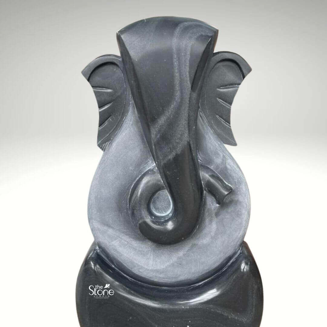 3 Feet Abstract Ganesha Statue 2 A striking three-foot stone abstract Ganesha statue meticulously crafted from black stone is presented against a neutral background emphasizing its modern artistry and symbolic form. This contemporary interpretation of Lord Ganesha, ideal for an article discussing abstract Ganesha sculptures features a minimalist yet recognizable representation of the beloved deity. The statue predominantly uses sweeping smooth curves and simplified forms to suggest Ganesha's iconic elephant head trunk, and ears rather than a literal depiction allowing for a more interpretive and artistic appreciation. The sculpture uniquely utilizes two contrasting tones of black stone creating a dynamic visual effect. The outer darker black stone forms the core outline of Ganesha's head and a sturdy rounded base. Within this, a lighter subtly textured gray black stone highlights the central features particularly defining the elongated trunk that elegantly curls to the left and the broad fan-like ears. The interplay of these two dark tones adds depth and sophistication to the abstract design allowing light to catch the varying surfaces and reveal subtle striations and natural patterns within the stone. The overall form is vertical and streamlined with the top suggesting Ganesha's crown and the bottom forming a stable elegant base. Despite its abstract nature the essence of Ganesha — wisdom, prosperity and the remover of obstacles — is clearly conveyed through the familiar contours and symbolic representation. This three-foot blackstone abstract Ganesha statue is a contemporary work of art that seamlessly blends spiritual symbolism with modern design making it a unique and powerful addition to any modern home office, or art collection as suggested by its context.