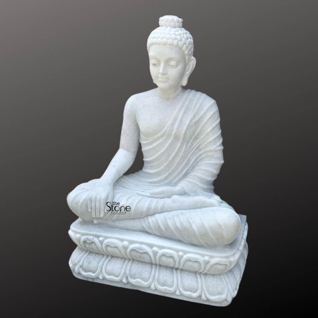3 Feet Marble Buddha Idol 2 A magnificent three-foot white marble Buddha statue ideal for a home setting is elegantly presented against a dark neutral background highlighting its pristine material and serene form. This exquisite statue depicts the Buddha seated in a meditative lotus position emanating an aura of profound peace enlightenment and spiritual tranquility making it a perfect addition to a sacred space within a home. His right hand rests gently on his knee in the Bhumisparsha Mudra symbolizing his moment of enlightenment under the Bodhi tree while his left hand rests gracefully in his lap palm facing upwards. The craftsmanship of the statue is exceptional showcasing the smooth luminous white marble that exquisitely captures the delicate details of the Buddha's form. The natural veining and subtle translucence of the marble enhance its artistic beauty. His robes are gracefully draped over his body with naturalistic flowing pleats that add depth and realism without detracting from the overall serene aesthetic. The Buddha's facial features are finely sculpted with a calm and contemplative expression characteristic of traditional Buddhist art, inviting introspection. A prominent ushnisha on his head symbolizes wisdom and spiritual attainment while his hair is meticulously carved into tight symmetrical curls. His elongated earlobes are indicative of his princely past and renunciation of worldly possessions. The Buddha is seated upon a beautifully carved lotus pedestal also made from pristine white marble. This pedestal features multiple layers of intricately detailed lotus petals symbolizing purity spiritual awakening and beauty. The luminous white marble softly reflects light, giving the statue a radiant quality that enhances its divine presence and makes it a striking focal point. This three-foot white marble Buddha statue is not merely a work of art but a powerful spiritual artifact, perfectly suited for a home temple meditation corner or a prominent place in a living space inspiring devotion inner peace and a sense of calm.