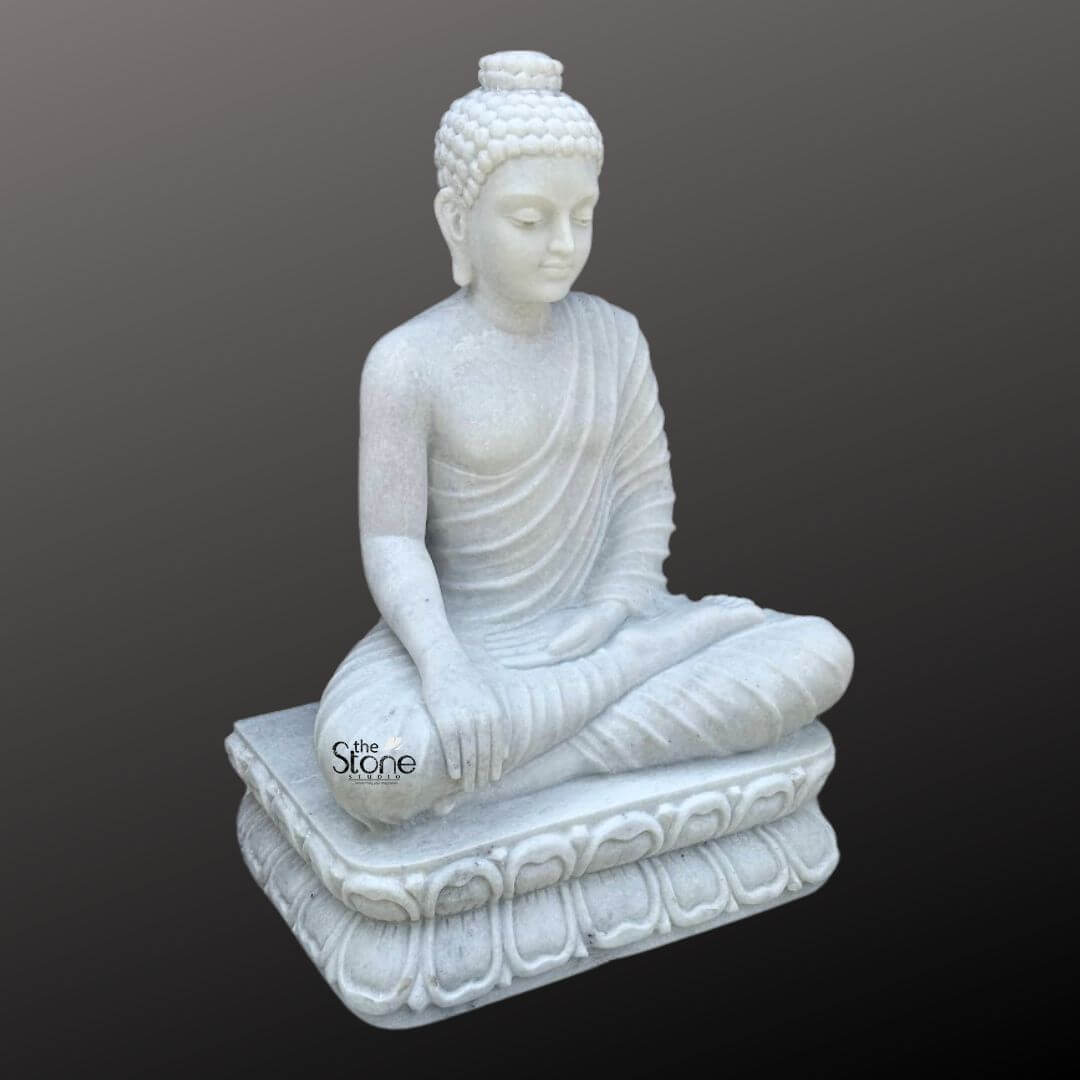 3 Feet Marble Buddha Idol 5 A magnificent three-foot hand-carved marble Buddha statue is elegantly presented against a dark neutral background highlighting its pristine white material and serene form. This exquisite statue central to an article about hand-carved marble Buddha statues depicts the Buddha seated in a meditative lotus position emanating an aura of profound peace enlightenment and spiritual tranquility. His right hand rests gently on his knee in the Bhumisparsha Mudra symbolizing his moment of enlightenment under the Bodhi tree while his left hand rests gracefully in his lap palm facing upwards. The craftsmanship of the hand-carved statue is exceptional showcasing the smooth luminous white marble that exquisitely captures the delicate details of the Buddha's form. The natural veining and subtle translucence inherent in the marble enhance its artistic beauty and unique character. His robes are gracefully draped over his body with naturalistic flowing pleats that add depth and realism without detracting from the overall serene aesthetic. The details of the folds are expertly carved suggesting soft fabric while being rendered in hard marble. The Buddha's facial features are finely sculpted with a calm and contemplative expression characteristic of traditional Buddhist art inviting introspection and reverence. A prominent ushnisha on his head symbolizes wisdom and spiritual attainment with his hair meticulously carved into tight symmetrical curls. His elongated earlobes are indicative of his princely past and renunciation of worldly possessions. The Buddha is seated upon a beautifully hand-carved lotus pedestal also made from pristine white marble. This pedestal features multiple layers of intricately detailed lotus petals symbolizing purity spiritual awakening and beauty. The luminous white marble softly reflects light giving the statue a radiant quality that enhances its divine presence and makes it a striking focal point. This three-foot hand-carved marble Buddha statue is not merely a work of art but a powerful spiritual artifact perfectly suited for a temple meditation center or a prominent place in a home inspiring devotion inner peace and a sense of calm.