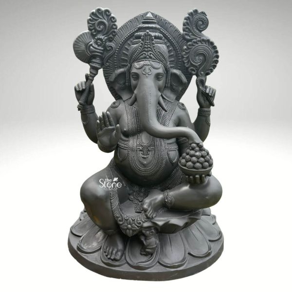 A magnificent 4-foot Ganesha statue crafted from fiber and finished to resemble dark stone is presented against a clean softly diffused background that transitions from light grey to white. Lord Ganesha is seated in a relaxed yet regal posture with his legs crossed resting upon a beautifully sculpted lotus pedestal. This large Ganesha statue is a striking representation of the beloved deity perfect for significant spaces. Ganesha is depicted with his iconic elephant head featuring large ears intricate detailing around his trunk and expressive eyes. His four arms hold various attributes: his upper right hand holds an axe or goad, and his upper left hand holds a noose symbolizing control and guidance. His lower right hand is raised in the Abhaya Mudra, a gesture of fearlessness and blessing, while his lower left hand rests on his knee holding a bowl filled with Modaks which his trunk playfully reaches towards. Behind Ganesha's head an elaborate arching prabhavali extends upwards featuring intricate scrollwork and divine motifs enhancing his divine presence. The statue's surface though made of fiber is meticulously textured to mimic the natural grain and appearance of dark stone, giving it a weighty and traditional aesthetic. Despite its substantial 4-foot size, the fiber material would make it relatively lighter than solid stone, offering practical advantages. This detailed and imposing 4 feet big Ganesha statue beautifully conveys prosperity, wisdom, and obstacle removal, directly aligning with an article discussing large-scale Ganesha sculptures.