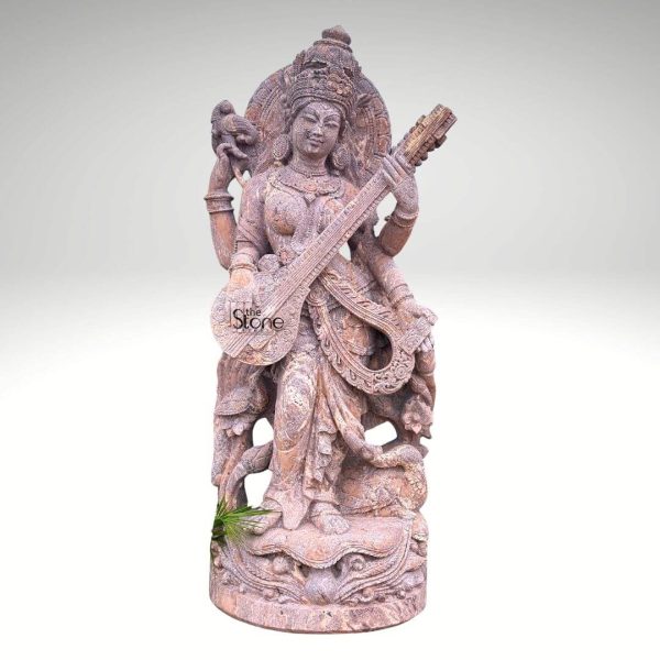 A magnificent 6-foot tall standing Saraswati statue meticulously carved from reddish-brown sandstone embodies the goddess of knowledge music art wisdom and nature. Goddess Saraswati is depicted gracefully standing playing her veena a stringed instrument with her two main hands holding a sacred text in one of her upper hands and a rosary in another symbolizing learning and meditation. Her serene face features downcast eyes and a gentle smile adorned with an elaborate crown and intricate jewelry including necklaces armlets and anklets. Her elegant form is draped in finely sculpted robes with exquisite folds and patterns enhancing her divine grace. Behind her a halo or prabhavali decorated with traditional motifs signifies her divine radiance. The statue features additional intricate carvings around its base which depicts natural elements like foliage and possibly her vahan the swan or peacock adding to the rich narrative. The natural texture and warm earthy tones of the sandstone lend an ancient and organic quality to the sculpture with subtle variations in color that enhance its natural beauty. A small green leaf or plant sprouts from the base on the left subtly integrating the statue with nature. Presented against a plain, light grey background the majestic height and intricate details of the sandstone Saraswati statue are beautifully highlighted. Its impressive 6-foot stature ensures it makes a commanding and deeply spiritual presence ideal for a temple an educational institution a grand interior space or a specialized spiritual garden serving as a powerful symbol of wisdom creativity and spiritual enlightenment.
