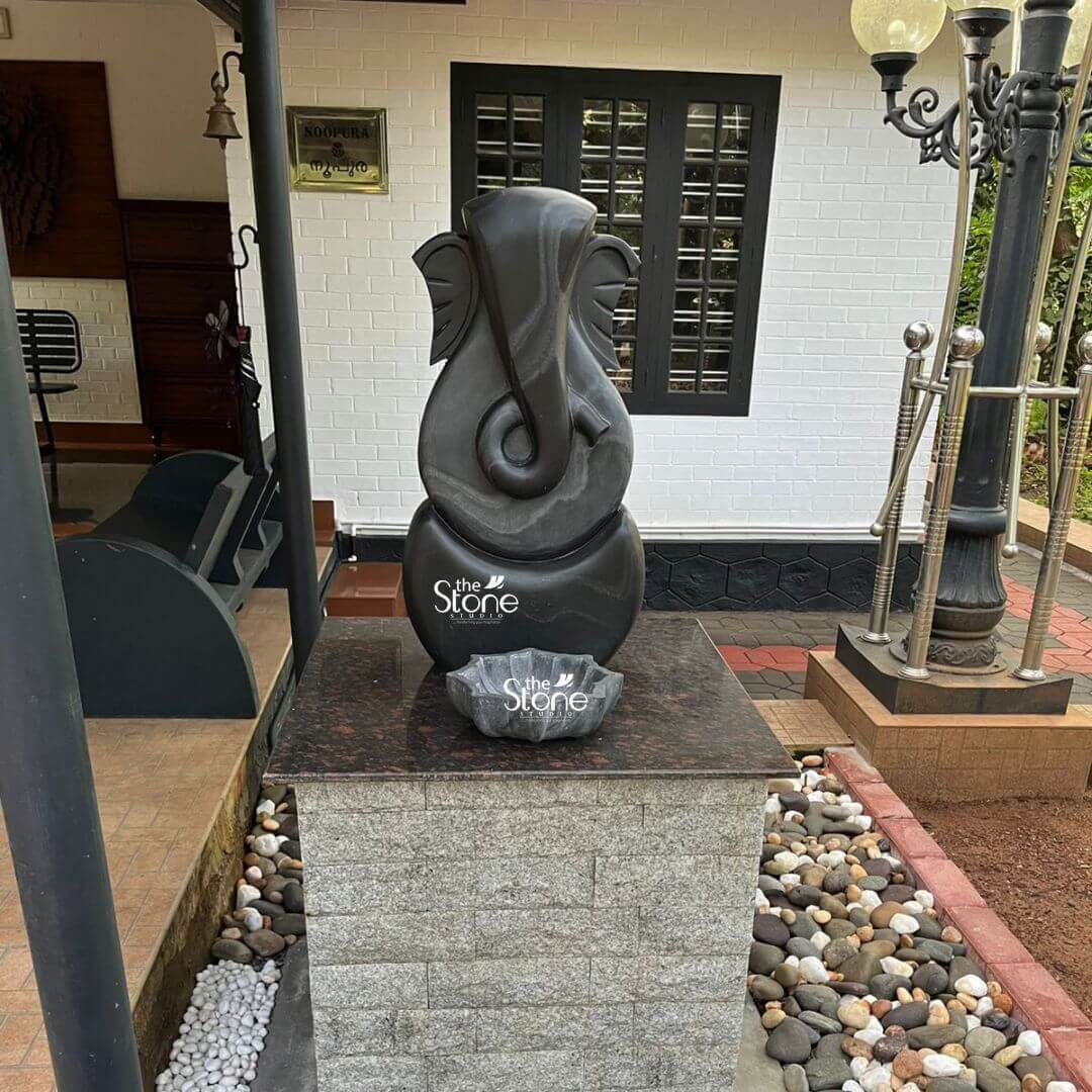 3 Feet Abstract Ganesha Statue 3 A striking three-foot black stone abstract Ganesha idol is prominently displayed on a granite-topped pedestal in a modern home's exterior or entrance area creating a contemporary spiritual focal point. This statue ideal for an article discussing abstract Ganesha idols for home features a minimalist yet recognizable representation of Lord Ganesha blending spiritual symbolism with modern design. The sculpture predominantly uses sweeping smooth curves and simplified forms to suggest Ganesha's iconic elephant head trunk and ears, rather than a literal depiction. The statue utilizes two contrasting tones of black stone creating a dynamic visual effect. The outer darker black stone forms the core outline of Ganesha's head and a rounded base. Within this, a lighter subtly textured gray-black stone highlights the central features particularly defining the elongated trunk that elegantly curls to the left and the broad fan-like ears. The interplay of these two dark tones adds depth and sophistication to the abstract design allowing natural light to catch the varying surfaces and reveal subtle striations in the stone. The Ganesha idol is positioned on a sturdy pedestal with a dark polished granite top and a base of rough-hewn light gray stone enhancing its presence. At the foot of the idol on the granite surface a small black stone bowl possibly for offerings or water adds another thoughtful detail. The base of the pedestal is surrounded by decorative pebbles in varying shades of white gray and brown creating an attractive border. In the background, a modern white brick house with black-framed windows and a dark porch swing contributes to the contemporary home setting. A traditional bell hangs near the entrance and an ornate black lamppost with steel accents stands to the right. This three-foot blackstone abstract Ganesha idol is a unique and powerful artistic statement perfectly suited for a modern home seeking both aesthetic appeal and spiritual significance.