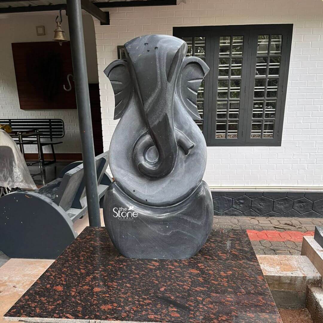 3 Feet Abstract Ganesha Statue 5 A striking three-foot black stone abstract Ganesha statue meticulously crafted for outdoor display is prominently situated on a dark red granite slab likely in a home's exterior or garden area. This statue ideal for an article discussing abstract Ganesha sculptures for outdoors features a minimalist yet recognizable representation of Lord Ganesha blending spiritual symbolism with modern design making it a versatile piece for various outdoor settings. The sculpture predominantly uses sweeping smooth curves and simplified forms to suggest Ganesha's iconic elephant head trunk, and ears rather than a literal depiction. The statue uniquely utilizes two contrasting tones of black stone creating a dynamic visual effect. The outer darker black stone forms the core outline of Ganesha's head and a sturdy rounded base. Within this, a lighter subtly textured gray-black stone highlights the central features particularly defining the elongated trunk that elegantly curls to the left and the broad fan-like ears. The interplay of these two dark tones adds depth and sophistication to the abstract design allowing natural light to catch the varying surfaces and reveal subtle striations and natural patterns within the stone. These inherent qualities of black stone ensure its durability and aesthetic appeal in outdoor environments. The Ganesha idol is positioned on a polished dark red granite slab with black and reddish speckles which serves as a sturdy and attractive base. The background features a modern white brick house with black-framed windows suggesting a contemporary residential setting. To the left, a dark outdoor bench and other garden elements are visible reinforcing the outdoor context. This three-foot blackstone abstract Ganesha statue is a contemporary work of art that seamlessly blends spiritual symbolism with modern design making it a unique and powerful addition to any garden, patio or outdoor spiritual space. Its robust material ensures it can withstand outdoor elements while maintaining its artistic integrity and serving as a remover of obstacles and bestower of prosperity.