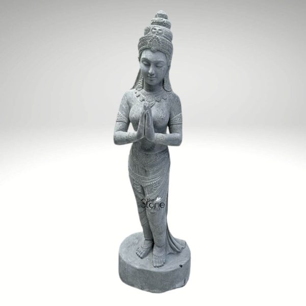 A graceful 6-foot tall Balinese Welcome Lady statue meticulously carved from black stone with a prominent greyish appearance stands on a circular base extending a traditional greeting. The figure is slender and elegantly proportioned with her hands pressed together at chest level in the Namaste or Anjali Mudra gesture symbolizing respect hospitality and reverence. She wears an elaborate tiered crown characteristic of Balinese or Southeast Asian deity sculptures beneath which her finely detailed hair cascades over her shoulders. Her body is adorned with intricate jewelry including a delicate necklace armbands and a detailed belt. Her lower body is draped in a finely sculpted sarong or traditional skirt featuring exquisite patterns and folds that enhance her elegant form. The natural texture and subtle variations in the greyish black stone are visible throughout the sculpture highlighting the intricate carvings of her attire jewelry, and serene facial features. Her eyes are gently closed or downcast conveying a sense of peacefulness and humility. The circular base provides a stable and dignified foundation for the tall slender figure. Presented against a plain light grey background the majestic height and intricate details of the Balinese Welcome Lady statue are beautifully accentuated. This sculpture is ideal for enhancing a home entrance garden, or any space desiring a touch of exotic elegance spiritual tranquility and the warm welcoming spirit of Balinese culture. Its impressive 6-foot stature ensures it makes a commanding and graceful statement.