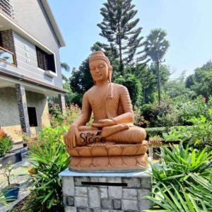 A magnificent 4-foot tall Buddha statue meticulously carved from reddish-brown sandstone is prominently displayed outdoors on an elevated stone pedestal ideal for a garden setting in Kolkata. The Buddha is seated in the Bhumisparsha Mudra with his right hand gently touching the ground symbolizing his enlightenment under the Bodhi tree and his calling the earth to witness his awakening. His left hand rests in his lap palm upward in a gesture of meditation. The Buddha's serene face features softly closed eyes a gentle smile and finely detailed features including his tightly coiled hair elongated earlobes, and the urna all conveying deep tranquility and spiritual wisdom. The flowing robes are intricately sculpted with delicate folds that emphasize his form. The natural texture and warm earthy tones of the sandstone lend an ancient and organic quality to the sculpture with subtle variations in color that enhance its natural beauty. The statue sits on a beautifully carved lotus pedestal symbolizing purity and spiritual growth. The elevated pedestal is constructed from grey and white stone blocks providing a sturdy and dignified foundation for the Buddha. The statue is surrounded by lush green plants and blooming flowers creating a vibrant and serene garden environment. In the background a modern house with grey and white exterior elements is visible blending contemporary architecture with traditional spiritual art. Tall trees and clear blue skies complete the peaceful outdoor scene. This image beautifully captures the spiritual essence and artistic grandeur of the Bhumisparsha Buddha making it an ideal focal point for a meditation garden or a peaceful outdoor sanctuary.
