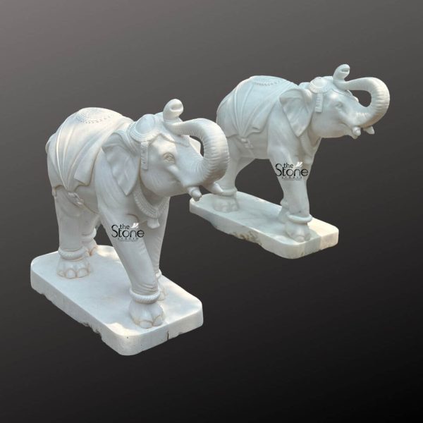 A majestic pair of four-foot tall elephant statues meticulously carved from white marble stand elegantly on matching rectangular bases. Each elephant is depicted with its trunk raised in a welcoming gesture and their bodies are adorned with intricate decorative carvings that simulate traditional ceremonial coverings. The detailing highlights their powerful forms from the gentle curve of their tusks to the texture of their skin and the strength of their legs. The statues convey a sense of grandeur and traditional craftsmanship making them ideal as decorative pieces or focal points. They are presented against a simple dark background which emphasizes their pristine white color and the exquisite artistry of the marble. These substantial elephant sculptures showcase exceptional artistry with each curve and line carefully considered to bring these magnificent creatures to life in stone. The smooth polished surface of the white marble reflects light enhancing the delicate etchings and patterns that mimic saddles, blankets and jewelry traditionally used in elephant adornment. From the detailed eyes to the powerful yet graceful, stance every element contributes to the statues realistic and regal presence. The symmetrical composition of the two elephants suggests a sense of balance and harmony making them perfect for grand entrances spacious gardens or as a commanding presence within large interior spaces. Their impressive scale at four feet makes them significant statement pieces, embodying strength, wisdom and good fortune qualities often associated with elephants. The robust bases provide stability and complete the statues dignified presentation ensuring they stand as enduring testaments to the beauty of natural stone and expert craftsmanship.