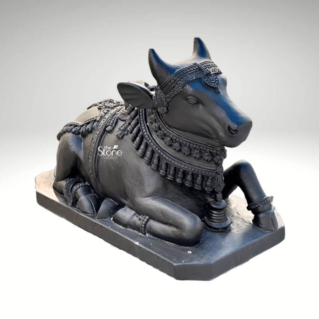 Black Nandi Bull Statue for Home 2.5ft 1 A majestic 2.5-foot black stone Nandi Bull statue designed as a sacred adornment for a home is prominently featured against a clean neutral background emphasizing its intricate details and serene presence. This exquisite sculpture depicts Nandi the revered vahana of Lord Shiva in a classic reclining posture symbolizing patience devotion and strength. The bull is shown lying down with its legs folded beneath its body its head gracefully turned slightly to the right looking forward. The craftsmanship of the statue is exceptional showcasing the smooth deep black stone that beautifully accentuates Nandi's powerful yet calm form. The bull is richly adorned with meticulously carved traditional Indian jewelry and ceremonial embellishments. These include an elaborate crown or headpiece on its forehead intricately designed necklaces with dangling ornaments around its neck and decorative blankets or saddle covers carved onto its back all integrated seamlessly into the stone. The details of the ornaments such as beads bells and patterns are finely rendered enhancing the statue's divine appearance. Nandi's facial features are finely sculpted with a calm and benevolent expression characterized by gentle eyes a strong snout and well-formed horns. The texture of the black stone with its inherent depth and subtle sheen gives the statue a timeless and dignified quality. The bull rests on a sturdy rectangular black stone base which provides stability and a formal presentation. This 2.5-foot blackstone Nandi Bull statue is not merely a decorative item but a powerful spiritual artifact, ideal for a home temple entrance or garden inspiring devotion and bringing an auspicious presence to the living space as highlighted in an article about such home decor.
