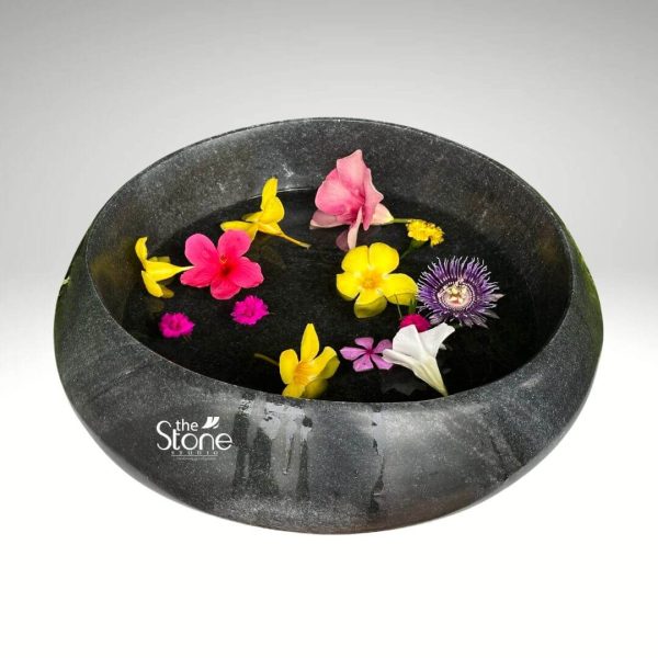 A beautiful black stone Urli is presented from a slightly elevated angle against a soft light gray background highlighting its elegance as a home decor item. The Urli is a shallow wide-mouthed bowl crafted from natural black stone with a subtly textured speckled finish that gives it a sophisticated earthy appeal. Its exterior is smoothly curved showcasing the organic beauty of the stone. The bowl is filled with clear water on the surface of which a vibrant array of fresh colorful flowers floats gracefully. The floral arrangement includes bright yellow hibiscus-like blooms rich pink hibiscus a striking purple passionflower and several smaller pink and white blossoms. These colorful elements create a striking contrast against the dark surface of the water and the black stone of the Urli making the display visually captivating. This traditional Urli design is perfect for enhancing home decor serving as a serene centerpiece for entryways living rooms or meditation spaces. The combination of the natural black stone and the floating flowers evokes a sense of tranquility and aesthetic beauty ideal for creating a calming and inviting atmosphere within a home. The craftsmanship of the black stone Urli suggests durability and a timeless appeal making it a lasting addition to any interior design scheme focused on natural materials and serene aesthetics.