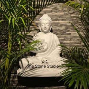 A serene 2-foot Buddha statue crafted from white fiber is beautifully showcased in a dimly lit tranquil setting, evoking a sense of calm and spiritual presence possibly in a home or garden in Madurai .The statue depicts Buddha seated in a traditional meditative posture likely the Dhyana Mudra with hands gently resting in his lap palms upwards. His eyes are softly closed conveying deep meditation inner peace and enlightenment. The fiber material gives the statue a pristine white, smooth, and slightly luminous finish allowing it to stand out elegantly against the darker background. Buddha's robes are finely sculpted with flowing realistic pleats suggesting soft fabric draped over his body. His head is adorned with tightly curled hair, an ushnisha at the crown signifying his spiritual wisdom, and elongated earlobes. His facial features are rendered with exquisite serenity, reflecting a profound inner tranquility. The statue rests on a simple, dark, cylindrical base, which provides a stable and aesthetically pleasing foundation. It is framed by lush, dark green foliage from tropical plants, possibly palm fronds, which create natural shadows and enhance the serene, contemplative atmosphere. In the background, a textured wall, possibly made of stone or brick, adds depth and a subtle, natural backdrop. The lighting creates a dramatic effect, highlighting the Buddha's form and enhancing the peaceful ambiance, making this fiber Buddha statue an ideal piece for a meditation space, garden, or indoor decor, inspiring calm and mindfulness.