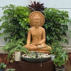 A serene 3-foot Buddha statue meticulously carved from sandstone is beautifully set in a lush outdoor garden designed to enhance a tranquil space. The statue depicts Buddha in a meditative seated posture with his hands in a mudra of concentration or teaching. His eyes are gently closed conveying a profound sense of inner peace and enlightenment. The sandstone material gives the statue a natural earthy brown hue with a subtly textured finish allowing it to blend harmoniously with the garden environment. Buddha's robes are finely sculpted with delicate pleats suggesting soft fabric. His head is adorned with tightly curled hair and an ushnisha at the crown signifies his spiritual wisdom. A prominent urna is visible on his forehead. Behind his head a beautifully carved circular halo or aura emanates featuring intricate traditional patterns that symbolize his divine wisdom and enlightenment. The statue rests on a dark, cylindrical pedestal. In front of the statue a wide dark green shallow bowl is filled with white pebbles and small succulents creating a miniature zen garden. Surrounding the Buddha are various vibrant green plants including ferns and other leafy foliage with a darker red leafed plant directly behind the halo adding depth and color to the natural setting. A light colored wall forms the backdrop contrasting with the dense greenery. This sandstone Buddha statue creates a peaceful and spiritual focal point perfect for a garden in Bangalore inviting contemplation and calm.