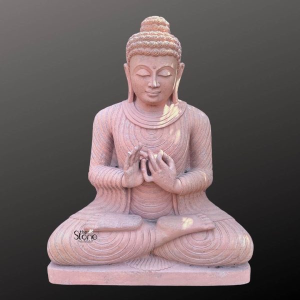 A majestic 3-foot tall Buddha statue meticulously carved from reddish sandstone sits in a profound meditative posture with his hands performing the Dharmachakra Mudra symbolizing the turning of the wheel of dharma or teaching. The Buddha's serene face features softly closed eyes a gentle smile and finely detailed features including his tightly coiled hair elongated earlobes and the urna all conveying deep tranquility and wisdom. The flowing robes are intricately sculpted with delicate concentric folds creating a sense of natural drapery that emphasizes his form. The Dharmachakra Mudra is clearly depicted with both hands held at chest level the right palm facing outwards and the left palm facing inwards with the thumbs and index fingers forming circles representing the continuous cycle of dharma. The natural texture and warm earthy tones of the reddish sandstone lend a unique inviting and ancient quality to the sculpture with subtle variations in color that enhance its organic beauty. The statue rests on a simple, sturdy base integral to its design providing a stable foundation. Presented against a plain dark grey background the warm hue and intricate details of the sandstone Buddha statue are beautifully highlighted. Its impressive 3-foot height makes it a significant focal point ideal for a meditation space, garden or as a central element in a spiritual sanctuary. This Dharmachakra Mudra Buddha statue serves as both a beautiful artistic element and a powerful symbol of teaching enlightenment and the path to spiritual awakening.