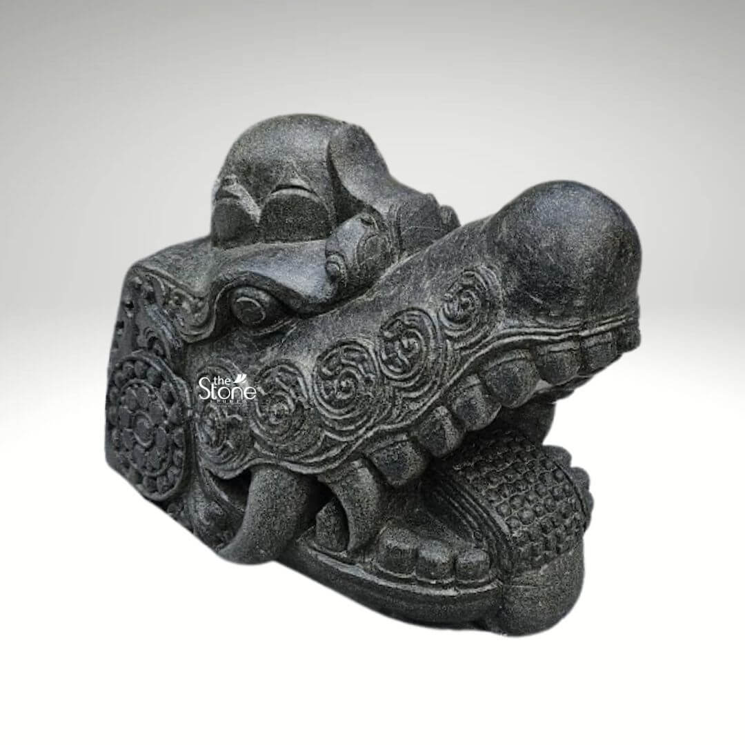 Dragon Head Statue 18" 6 A detailed black stone dragon head idol measuring 1.5 feet in length and 1 foot in height is expertly carved and displayed against a plain light background. This sculpture represents a powerful and revered mythical creature perfect for an article discussing dragon head idols. The dragon's head is rendered with striking realism and artistic flair featuring a robust snout and a wide-open mouth that reveals formidable stylized fangs and a meticulously textured interior conveying an impression of immense power and ancient wisdom. The rich dark black stone material gives the idol a profound and timeless quality emphasizing its strength and durability. The entire surface of the dragon's head is adorned with an array of intricate traditional carvings. These include flowing swirling patterns that simulate the complex texture of scales and suggest dynamic movement along with prominent circular motifs. These circular designs reminiscent of ancient symbols or stylized floral elements are strategically integrated into the overall ornamentation adding significant cultural depth and artistic elegance to the piece. The eyes are deeply set and carved with a piercing vigilant gaze enhancing the idol's commanding and protective demeanor. A distinctive crown-like or horn-like protrusion on the top of the head further accentuates its majestic and mythical status reinforcing its symbolic power as a revered idol. The craftsmanship is exemplary showcasing varied textures across the sculpture from the smoother polished areas to the more rugged detailed scales and ridges along the jawline and brow. This intricate play of textures creates a dynamic visual effect allowing light and shadow to highlight the three-dimensional form and bring the mythical creature to vivid life. This blackstone dragon head idol is more than just a decorative item it serves as a powerful cultural and spiritual artifact embodying strength good fortune and protection. Its size and material make it suitable for a prominent display whether in a private collection, a public space, or a dedicated shrine as implied by its description as an idol.