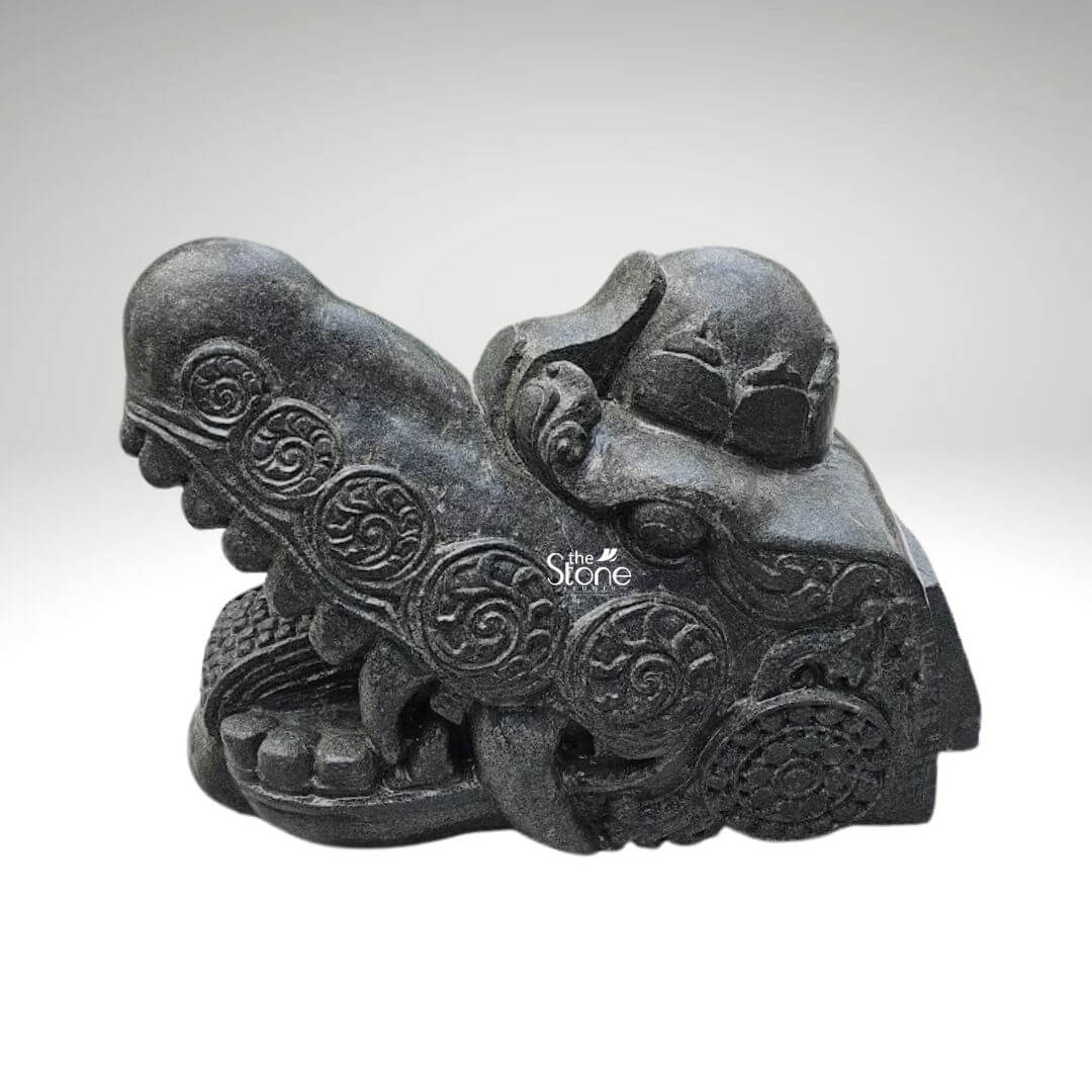 Dragon Head Statue 18" 7 A robust black stone dragon head sculpture measuring 1.5 feet in length and 1 foot in height is showcased against a soft, light gray background highlighting its intricate details and suitability as a striking garden feature. This meticulously carved statue embodies the mythical power and ancient wisdom of a dragon designed to add a dramatic and cultural element to an outdoor setting. The dragon's head is depicted with a formidable snout and a wide-open mouth revealing sharp stylized fangs and a deeply textured interior conveying an impression of vigilance and strength. The dark rich black stone material gives the sculpture a timeless and weathered appearance perfectly suited to blend with natural garden elements while standing out as a focal point. The entire surface of the dragon's head is adorned with an array of traditional flowing carved patterns including swirling motifs that mimic scales and suggest dynamic energy. Prominent circular designs reminiscent of ancient symbols or stylized floral elements are strategically integrated into the ornamentation adding layers of cultural depth and aesthetic appeal. The eyes are deeply set and carved with a piercing, watchful gaze which enhances the sculpture's protective and majestic demeanor. A distinctive crown-like or horn-like protrusion on the top of the head further accentuates its mythical status reinforcing its symbolic power within a garden landscape. The craftsmanship is exemplary showcasing varied textures across the sculpture from the smoother more refined areas to the rugged detailed scales and ridges along the jawline and brow. This intricate play of textures creates a dynamic visual effect allowing natural light and shadow in a garden setting to highlight the three dimensional form and bring the mythical creature to vivid life. This blackstone dragon head sculpture is more than just a decorative item it serves as a powerful artistic statement symbolizing strength good fortune and guardianship making it an ideal enhancement for a garden as suggested by its context in an article about garden sculptures.