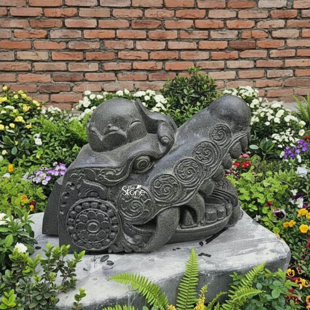 Dragon Head Statue 18" 3 A majestic black stone dragon head statue measuring 1.5 feet in length and 1 foot in height is strategically placed atop a large flat gray stone, nestled within a vibrant garden setting against a rustic brick wall. This sculpture is specifically designed to serve as a formidable and symbolic guardian for a gate or entrance embodying traditional protective qualities. The dragon's head is rendered with a powerful open snout, revealing sharp stylized fangs and a meticulously textured interior conveying an impression of alertness and strength ready to deter unwelcome energies. The dark rich black stone material gives the sculpture an ancient and robust quality perfectly suited for an outdoor architectural feature. The entire surface of the dragon's head is richly adorned with intricate traditional carvings including flowing swirling patterns that simulate scales and evoke dynamic movement along with prominent circular motifs. These circular designs reminiscent of ancient symbols or stylized floral patterns are strategically integrated into the ornamentation adding layers of cultural depth and aesthetic appeal. The eyes are deeply set and carved with a piercing watchful gaze which enhances the sculpture's commanding and vigilant demeanor. A distinctive crown-like or horn-like protrusion on the top of the head further accentuates its majestic and mythical status reinforcing its symbolic power as a gate guardian. The craftsmanship is exemplary showcasing varied textures across the sculpture from the smoother more refined areas to the rugged detailed scales and ridges along the jawline and brow. This intricate play of textures creates a dynamic visual effect allowing natural light and shadow to highlight the three dimensional form and bring the mythical creature to vivid life. The surrounding garden is lush with a variety of green plants and colorful flowers including white yellow, purple and red blooms creating a vibrant backdrop that contrasts beautifully with the dark stone of the dragon. The weathered brick wall in the background adds to the timeless and grounded feel of the scene. This blackstone dragon head sculpture is more than just a decorative item it serves as a powerful artistic and symbolic statement representing strength good fortune, and protection making it an ideal enhancement for a gate as suggested by its context in an article about such architectural features.