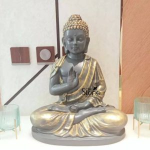 A captivating 1.5-foot tall Buddha statue meticulously crafted from fiber-reinforced plastic sits in a serene meditative posture with its hands in the Vitarka Mudra symbolizing teaching and intellectual discussion. The statue features a striking two-tone finish: the Buddha's body, face and hands are painted in a dark grey or charcoal color while his flowing robes and the textured curls of his hair are finished in a shimmering gold creating a rich and elegant contrast. His tranquil expression tightly coiled hair and elongated earlobes are finely detailed conveying deep peace and enlightenment. The statue rests on a simple flat base integral to its design and is prominently displayed indoors on a polished surface likely a table or console. It is flanked by two stylish ribbed clear glass candle holders or decorative cups on slender gold stands adding to the sophisticated ambiance. The background features a modern wall with abstract geometric patterns and two square decorative elements one dark and one with a circular cutout suggesting a contemporary interior design. This image beautifully showcases a versatile FRP Buddha statue as a focal point for home decor in places like Chennai blending traditional spiritual symbolism with modern aesthetic appeal. Its manageable size and unique finish make it an ideal decorative piece for shelves altars or side tables bringing an aura of tranquility wisdom and sophisticated elegance to any living space.