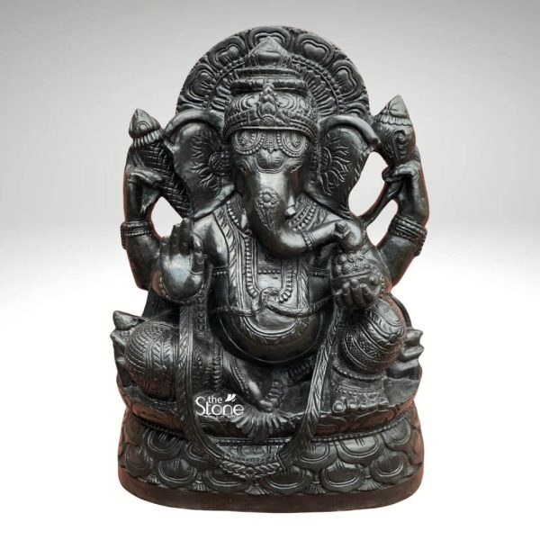 A captivating 20-inch Ganesha statue meticulously carved from black stone is prominently displayed against a clean, plain white background highlighting its suitability as a welcoming focal point for a foyer. The statue depicts Lord Ganesha seated in a traditional and benevolent posture likely the Lalitasana one leg tucked one hanging down or a similar relaxed pose embodying wisdom prosperity and auspiciousness. The black stone material gives the sculpture a rich deep charcoal gray hue with subtle natural speckles and a smooth polished finish that beautifully catches the light emphasizing the intricate details. Ganesha is lavishly adorned with elaborate finely carved details representing traditional jewelry and attire. These include a magnificent multi-tiered crown with intricate patterns ornate necklaces armlets and anklets all seamlessly integrated into the stone. His four arms are clearly visible: one hand is raised in a gesture of blessing another holds a modak with his trunk curving towards it while his upper hands hold his traditional attributes like an ankusha and a pasha. His large ears and expressive eyes convey wisdom and warmth. A prominent urna is visible on his forehead. The entire figure rests on a beautifully carved lotus pedestal also made of black stone with delicate petal designs providing a stable and aesthetically pleasing foundation. Behind his head an elaborate arched halo or prabhavali frames his figure featuring traditional motifs that symbolize his divine radiance and wisdom. This black stone Ganesha statue with its significant presence and intricate craftsmanship is an ideal piece to greet guests and invoke blessings in a home foyer combining spiritual significance with artistic elegance.