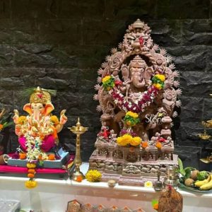A magnificent three-foot pink stone Ganesha statue is central to a beautifully decorated shrine likely in a home temple in Pune surrounded by offerings and another Ganesha idol creating a vibrant scene of devotion. This large Ganesha statue is intricately carved from pink stone giving it a warm earthy tone that stands out against the dark textured stone wall background. The deity is depicted in a traditional seated posture within an elaborate arch-like prabhavali that is also intricately carved with numerous small Ganesha figures and traditional motifs signifying his divine entourage and omnipotence. The main Ganesha figure is richly adorned with fresh garlands of yellow and red flowers draped gracefully around his neck and crown. He holds his traditional attributes in his multiple hands including a pasha ankusha and a modak. His trunk curls towards the left touching the modak. The details of his crown jewelry and robes are exquisitely carved showcasing the artisan's skill. The base of the pink stone statue is multi-tiered and features more intricate carvings including depictions of smaller deities or devotees further enhancing its grandeur. To the left of the large pink stone Ganesha, a smaller brightly colored Ganesha idol is placed on a raised platform adorned with fresh garlands of yellow and white flowers. This idol likely made of clay or another painted material contrasts in style and material with the main stone statue adding to the richness of the shrine. In front of the larger statue a traditional brass oil lamp and various puja offerings including bananas coconuts and other fruits are neatly arranged on a white shelf. The dark rough-hewn stone wall in the background provides a dramatic and authentic backdrop for the vibrant shrine. This entire composition evokes a deep sense of spirituality and celebration perfectly illustrating an article about a Ganesha stone statue in Pune showcasing its cultural and religious significance.