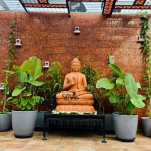 A serene 4-foot tall Buddha statue meticulously carved from reddish-brown sandstone is prominently featured as a focal point in an elegant outdoor garden or patio setting ideal for Bengaluru. The Buddha is seated in a meditative posture with his right hand raised in the Abhaya Mudra while his left hand rests gently in his lap. His face features a tranquil expression with softly closed eyes and his tightly coiled hair elongated earlobes and flowing robes are rendered with exquisite detail highlighting the natural texture and warm tones of the sandstone. The Buddha statue rests on a beautifully carved lotus pedestal which in turn is placed on a decorative black metal table with ornate legs. This table sits within a bed of green plants including tall bamboo-like foliage directly behind the statue and largeleafed plants in elegant grey planters flanking the table. The backdrop is a rustic red brick wall, possibly made of laterite stone adorned with small white decorative niches and climbing green vines that reach up towards a stylish pergola overhead. The entire scene creates a peaceful and harmonious ambiance blending spiritual tranquility with natural beauty and architectural elements. The stone paving in the foreground completes the sophisticated outdoor living space. This image beautifully captures how a magnificent sandstone Buddha statue can enhance a garden or patio inviting contemplation serenity and a touch of spiritual elegance to any home.