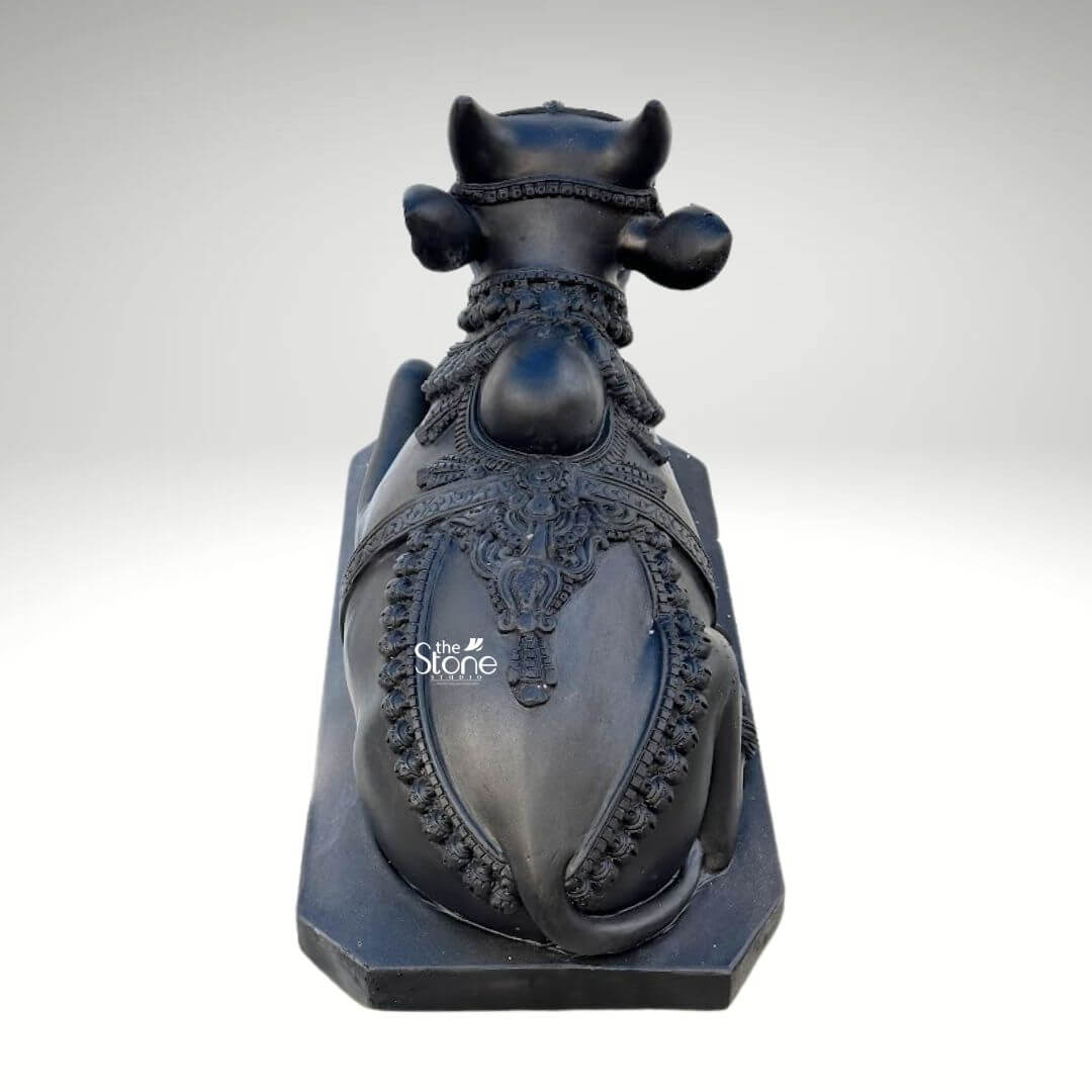 Black Nandi Bull Statue for Home 2.5ft 8 A majestic 2.5-foot hand-carved black stone Nandi statue specifically crafted for a Lord Shiva Mandir is prominently featured from a slightly elevated rear-side perspective against a clean neutral background emphasizing its intricate details and sacred presence. This exquisite sculpture depicts Nandi the revered vahana and eternal devotee of Lord Shiva in a classic reclining posture symbolizing unwavering patience devotion and strength making it ideal for an article discussing hand-carved Nandi statues for Shiva Mandirs. The bull is shown lying down with its legs gracefully folded beneath its body its head turned slightly to the right looking forward with a calm and benevolent expression. The craftsmanship of the hand-carved statue is exceptional showcasing the smooth deep black stone that beautifully accentuates Nandi's powerful yet serene form. The bull is richly adorned with meticulously carved traditional Indian jewelry and ceremonial embellishments. These include an elaborate crown or headpiece on its forehead intricately designed necklaces with dangling ornaments around its neck and decorative blankets or saddle covers carved onto its back all integrated seamlessly into the stone. The details of the ornaments, such as beads bells, and elaborate patterns, are finely rendered enhancing the statue's divine appearance and cultural authenticity highlighting the artisan's skill. From this view the intricate patterns on Nandi's back and sides outlined by decorative borders are particularly prominent emphasizing the elaborate craftsmanship. Nandi's well-formed horns rise gracefully from his head and his large, expressive ears are visible. The texture of the black stone, with its inherent depth and subtle sheen gives the statue a timeless and dignified quality, suitable for veneration in a sacred space. The bull rests on a sturdy, rectangular black stone base, which provides stability and a formal presentation consistent with an object of worship. This 2.5-foot hand-carved blackstone Nandi statue is not merely a decorative item but a powerful spiritual artifact inspiring deep devotion and bringing an auspicious presence to any Lord Shiva Mandir.