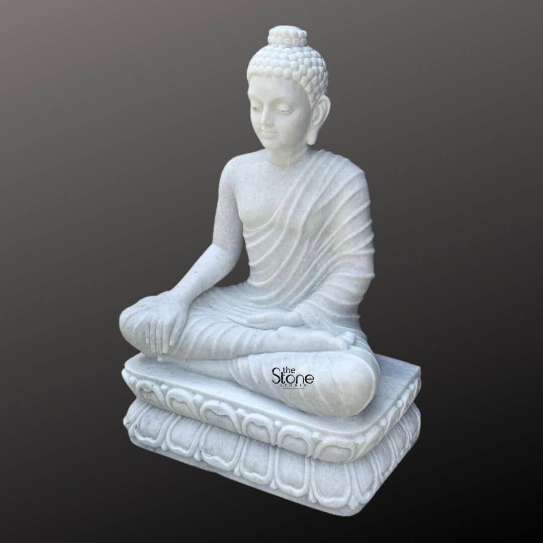 3 Feet Marble Buddha Idol 7 A magnificent three-foot handcrafted marble Buddha sculpture is elegantly presented against a dark neutral background highlighting its pristine white material and serene form. This exquisite statue central to an article about handcrafted marble Buddha sculptures depicts the Buddha seated in a meditative lotus position emanating an aura of profound peace enlightenment and spiritual tranquility. His right hand rests gently on his knee in the Bhumisparsha Mudra symbolizing his moment of enlightenment under the Bodhi tree while his left hand rests gracefully in his lap palm facing upwards. The craftsmanship of the handcrafted sculpture is exceptional showcasing the smooth luminous white marble that exquisitely captures the delicate details of the Buddha's form. The natural veining and subtle translucence inherent in the marble enhance its artistic beauty and unique character. His robes are gracefully draped over his body with naturalistic flowing pleats that add depth and realism without detracting from the overall serene aesthetic. The details of the folds are expertly carved suggesting soft fabric while being rendered in hard marble. The Buddha's facial features are finely sculpted with a calm and contemplative expression characteristic of traditional Buddhist art inviting introspection and reverence. A prominent ushnisha on his head symbolizes wisdom and spiritual attainment with his hair meticulously carved into tight symmetrical curls. His elongated earlobes are indicative of his princely past and renunciation of worldly possessions. The Buddha is seated upon a beautifully handcrafted lotus pedestal, also made from pristine white marble. This pedestal features multiple layers of intricately detailed lotus petals symbolizing purity spiritual awakening and beauty. The luminous white marble softly reflects light, giving the statue a radiant quality that enhances its divine presence and makes it a striking focal point. This three-foot handcrafted marble Buddha sculpture is not merely a work of art but a powerful spiritual artifact perfectly suited for a temple meditation center or a prominent place in a home inspiring devotion inner peace and a sense of calm.