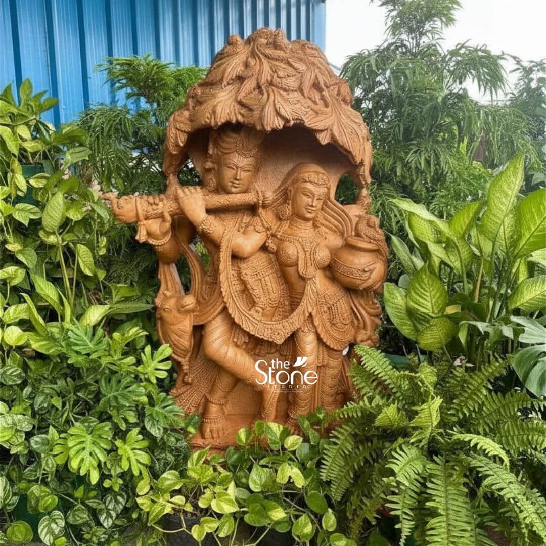 Radha Krishna Idol for Garden Décor 3.5ft 3 A captivating 3.5-foot Krishna Radha Murti meticulously carved from brown sandstone is beautifully nestled amidst a lush vibrant green garden enhancing its natural charm. The divine couple, Radha and Krishna are depicted standing together under an intricately sculpted tree canopy, with Krishna playing his enchanting flute. Lord Krishna stands gracefully on the left holding a flute to his lips, captivating Radha with his divine music. He is adorned with an elaborate crown traditional jewelry and flowing garments all finely detailed in the sandstone. A peacock an iconic symbol associated with Krishna is intricately carved near his feet further enriching the scene. Radha stands beside him on the right her expression one of deep devotion and enchantment holding a pot or pitcher symbolizing offerings or the abundance of nature. She too is adorned with a crown, necklaces armlets, and a flowing skirt all rendered with exquisite detail. The most prominent feature is the overarching tree canopy above them with its dense, realistic foliage carved in high relief from the same brown sandstone, providing a serene and naturalistic shelter. The entire 3.5-foot Krishna Radha Murti rests on a sturdy, multi-tiered rectangular base adorned with traditional decorative motifs, though partially obscured by the surrounding plants. The warm earthy tone and subtle texture of the brown sandstone give the sculpture an organic feel, allowing it to blend seamlessly with the outdoor garden environment. In the background, hints of a blue structure suggest an outdoor or semi-outdoor setting. This detailed brown sandstone sculpture beautifully embodies love, devotion, and harmony, directly aligning with an article discussing Krishna Radha Murti with a flute under a tree, perfect for garden settings.