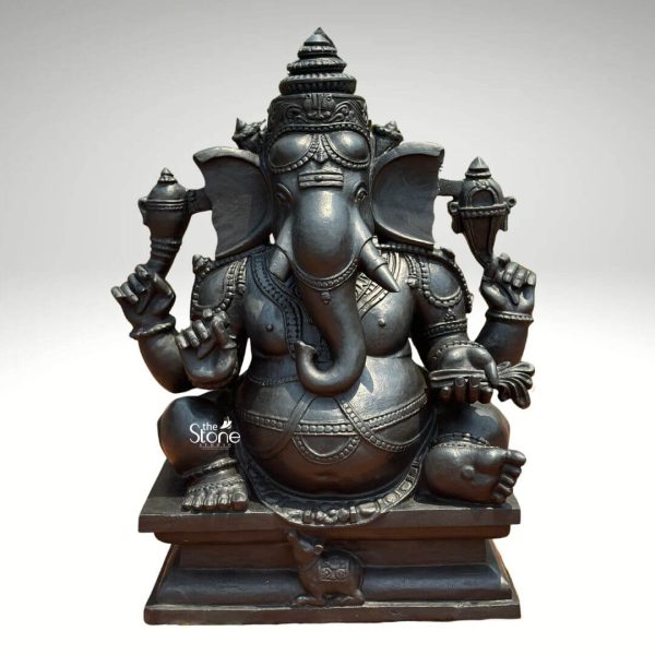 A magnificent three-foot black stone Kshipra Ganapati statue is presented against a neutral background highlighting its intricate details and serene presence. The deity Ganesha is depicted in a seated posture radiating an aura of wisdom and auspiciousness. Kshipra Ganapati known for quickly granting boons is shown with four arms each holding symbolic attributes. His upper right hand gracefully holds a pasha while his upper left hand grasps an ankusha representing control over desires and obstacles. His lower right hand holds his broken tusk symbolizing sacrifice and wisdom while his lower left hand offers a pot of modak indicating abundance and blessings. The sculpture's craftsmanship is evident in the smooth dark black stone which accentuates the round benevolent form of Ganesha. He is adorned with elaborate jewelry, including a regal crown intricate necklaces armlets, and anklets all meticulously carved into the stone. His large ears are beautifully fanned and his gentle eyes beneath arched brows convey a sense of calm and insight. The characteristic large belly of Ganesha is prominently displayed symbolizing prosperity and the ability to consume all sorrows. His trunk curves gracefully towards his left touching the modak pot, a classic depiction. Ganesha is seated on a simple yet sturdy square pedestal which is also carved from black stone. At the base of the pedestal his faithful vahana a tiny mouse is charmingly depicted looking up at the deity. The overall impression of the statue is one of reverence tradition, and profound spirituality making it a perfect subject for an article about Kshipra Ganapati. The black stone finish adds to its timeless appeal and traditional aesthetic, suitable for temples homes, or spiritual centers.