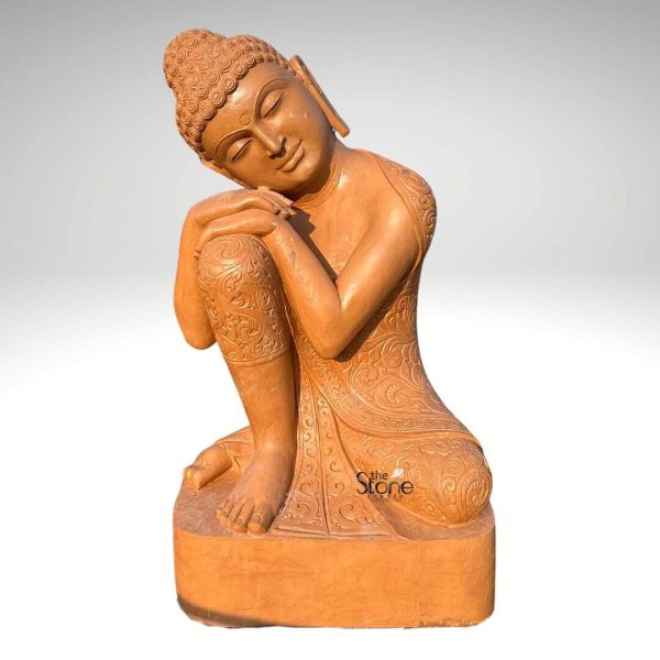 A large 6-foot Buddha statue exquisitely carved from sandstone is presented from a slightly elevated angle against a clean plain white background highlighting its unique contemplative posture and intricate details. The statue depicts Buddha in a seated somewhat curled-up position with his head gently resting on his raised knees or arms conveying a profound sense of introspection deep meditation and peaceful slumber or rest. His eyes are softly closed and a serene smile graces his lips. The sandstone material gives the sculpture a beautiful natural earthy reddish-brown hue with subtle textures emphasizing its organic origins and handcrafted artistry. Buddha's robes are intricately detailed with fine etched patterns resembling delicate floral or traditional motifs, especially visible on his legs and torso adding a rich ornamental quality. His head is adorned with tightly curled hair an ushnisha at the crown signifying his spiritual wisdom and elongated earlobes. The entire figure rests on a simple circular or oval sandstone base which provides a stable and aesthetically pleasing foundation. This posture distinct from typical meditative or reclining forms suggests a moment of profound personal reflection or a dream-like state making it a unique and captivating piece. The statue's significant height and natural stone composition make it a durable and timeless work of art ideal for a prominent display in a garden meditation space or large interior inspiring peace contemplation, and spiritual depth through its striking pose and intricate craftsmanship.