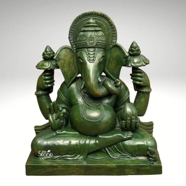 A captivating 21-inch tall Lord Ganesha statue meticulously carved from rich greenstone is depicted seated in a traditional cross-legged posture emanating an aura of wisdom and prosperity. The deity's iconic elephant head features large fanned ears and a gracefully curved trunk holding a modak in one of his hands symbolizing abundance. His four hands hold various attributes: two upper hands grasp lotus flowers one lower hand is raised in a gesture of blessing and the fourth hand holds the modak Lord Ganesha is adorned with intricate jewelry including necklaces and armlets and wears a beautifully detailed crown. A halo or prabhavali with radiating patterns frames his head signifying his divine radiance. The greenstone material gives the statue a unique earthy, and vibrant hue with subtle variations in color and texture that add to its natural beauty and depth. The polished surface catches the light highlighting the intricate carvings of his robes and ornaments. The statue rests on a simple sturdy rectangular base providing a solid foundation for this revered idol. Presented against a plain, light grey background the rich green color and detailed craftsmanship of the greenstone statue are beautifully accentuated. This depiction of Lord Ganesha is culturally significant representing new beginnings success, and the removal of obstacles, making it an ideal focal point for worship and decoration in homes or temples in places like Betul, Madhya Pradesh. Its moderate size makes it versatile for various placement options.