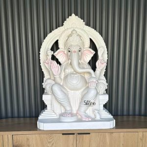 A striking 3-foot fiber Ganesha statue is presented front and center seated majestically on a light wooden surface with a contemporary dark gray ribbed wall as its backdrop. The statue portrays Lord Ganesha in a regal relaxed posture with his legs crossed at the ankles and his four arms holding traditional attributes while one hand is raised in a gesture of blessing. The entire statue is rendered in a pristine white or off-white finish giving it a pure serene, and elegant aesthetic. Subtle yet distinct pink accents are meticulously applied to Ganesha's palms, the soles of his feet and the inner part of his large expressive ears providing a gentle contrast and adding a touch of vibrancy to the otherwise monochromatic figure. His trunk gracefully curves to the left, and a traditional red tilak mark adorns his forehead signifying spiritual significance. Ganesha's eyes are open and gaze directly forward conveying wisdom benevolence, and calm. He is adorned with an intricately detailed crown necklaces and armlets all exquisitely sculpted into the fiber material. A magnificent arched halo or prabhavali frames his back featuring elaborate traditional carvings that enhance his divine presence. The statue rests upon a multi-tiered pedestal, also in white elevating the figure and adding to its grandeur. To the right of Ganesha's feet, a small white sculpted mouse his traditional vehicle (vahana), sits humbly, adding an iconic detail. This exquisite fiber Lord Ganesha statue serves as a powerful and beautiful focal point ideal for a home or spiritual space in New Zealand blending cultural heritage with artistic craftsmanship.