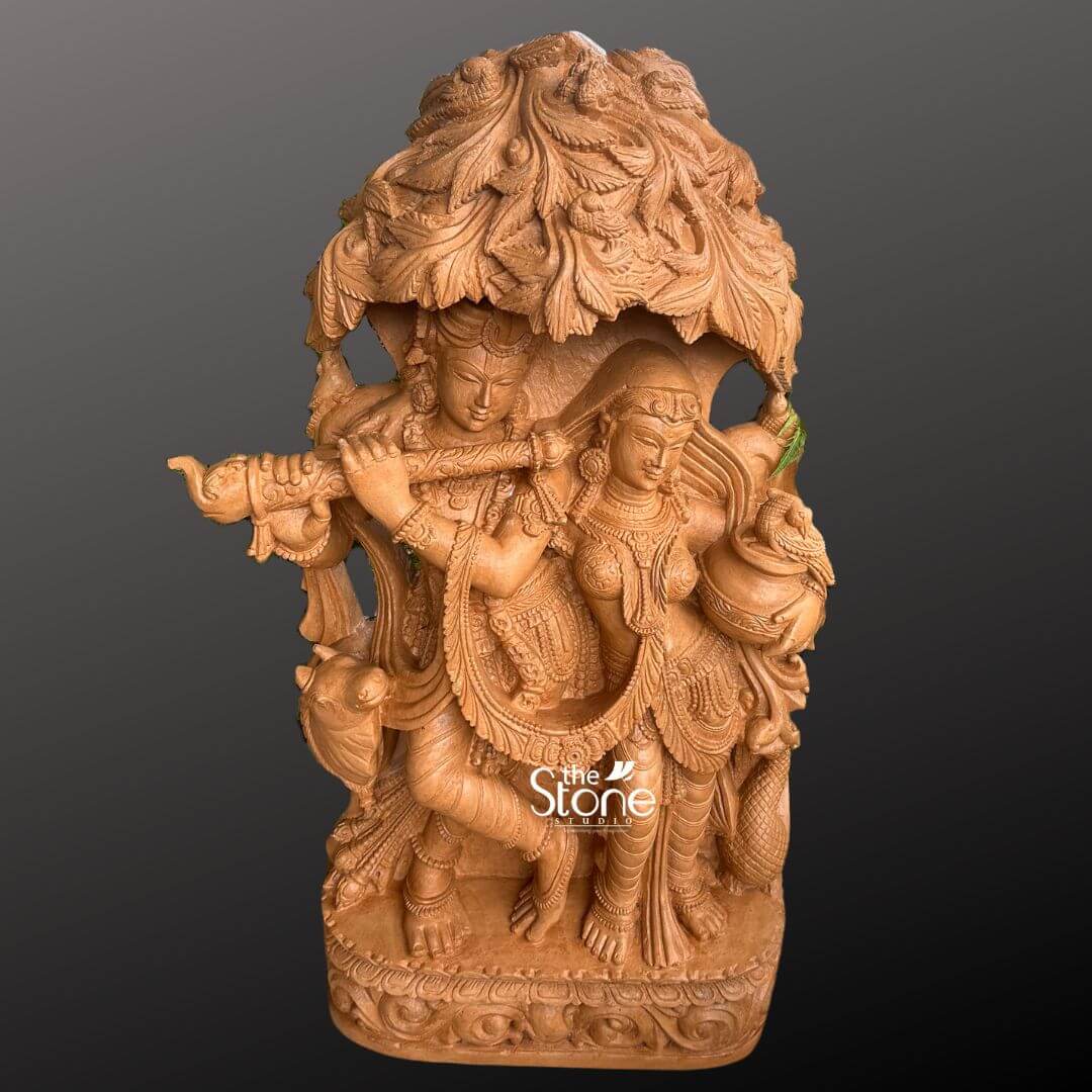 Radha Krishna Idol for Garden Décor 3.5ft 2 A magnificent 3.5-foot Lord Krishna playing flute with Radha statue exquisitely carved from brown sandstone is prominently displayed against a deep dark grey background highlighting its intricate details. The divine couple Radha and Krishna, are depicted standing gracefully together under an elaborate intricately carved tree canopy, with Lord Krishna serenading Radha with his flute. Lord Krishna stands on the left holding a flute to his lips his expression one of divine charm as he plays. He is adorned with an ornate crown, elaborate necklaces, armlets, and flowing dhoti all rendered with exceptional detail in the brown sandstone. A peacock a cherished symbol associated with Krishna is intricately carved near his feet adding a traditional element to the scene. Radha stands gracefully beside him on the right, her face expressing devotion and enchantment, holding a pot or pitcher symbolizing her offerings or connection to nature's bounty. She is also beautifully adorned with a crown necklaces, and a flowing sari with all drapery and jewelry finely sculpted. The most striking feature is the dense overarching tree canopy above them meticulously carved with numerous leaves and branches in high relief from the same brown sandstone providing a naturalistic and sacred shelter. The entire 3.5-foot Lord Krishna playing flute with Radha statue rests on a sturdy multi-tiered rectangular base which is richly decorated with traditional scrolling and floral motifs along its edges. The warm, earthy tone and subtle texture of the brown sandstone give the sculpture a natural and timeless aesthetic. This detailed brown sandstone sculpture beautifully embodies eternal love devotion, and the enchanting essence of Lord Krishna playing flute with Radha directly aligning with an article discussing such a revered idol.