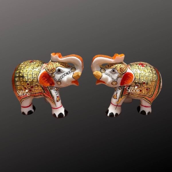 A pair of exquisite small marble elephant figurines are displayed side-by-side facing each other in a mirror-like pose against a plain dark gray background. Each elephant is intricately decorated showcasing traditional Indian artistry often associated with marble craftsmanship. The base material of the figurines is pristine white marble providing a clean and elegant canvas for the vibrant hand-painted details. Each elephant is richly adorned with golden red, and orange patterns that cover their bodies mimicking elaborate ceremonial coverings. The patterns include intricate geometric designs floral motifs and what appear to be small, embedded red beads or faux jewels, adding texture and sparkle. Gold paint highlights their ears trunks and the edges of their decorative blankets. Their faces are delicately painted with expressive eyes long eyelashes and ceremonial markings on their foreheads and cheeks. A string of tiny white beads is painted around their trunks and faces simulating intricate jewelry. Their large fanned ears are outlined in gold and feature a prominent orange-red patch on the inner side. The tips of their trunks and the hooves of their feet are painted black, providing a striking contrast to the white marble and rich colors. Both elephants have their trunks slightly raised a gesture traditionally associated with good luck and welcoming. These marble elephant figurines are perfect for adding a touch of cultural elegance and charm to home decor serving as symbols of strength wisdom and good fortune.
