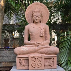 A serene 4-foot Meditating Buddha statue exquisitely carved from brown sandstone is presented outdoors against a soft-focus background of lush green foliage and architectural elements likely within a garden setting in Mumbai. The Buddha is depicted in a deep meditative posture seated with legs crossed in Vajrasana and hands gently resting in his lap in the Dhyana Mudra. The statue's light brown sandstone material gives it a warm, earthy tone and a subtle texture highlighting the intricate details. His face conveys profound peace with downcast eyes a gentle smile and elongated earlobes. His hair is composed of tight uniform curls, and an Ushnisha signifies his wisdom. A prominent circular halo intricately carved with radiating patterns is positioned behind his head symbolizing enlightenment and divine aura. The flowing drapery of his monastic robe is elegantly sculpted creating soft naturalistic folds. The Buddha sits on a square base which is also carved from brown sandstone and features significant Buddhist symbols. The front of the base displays a Dharma Chakra in the center, flanked by carved lions on either side, representing royalty, strength, and the spread of Buddhist teachings. The entire 4-foot meditating Buddha stone statue stands on a slightly elevated stone platform further enhancing its majestic presence. This detailed brown sandstone sculpture beautifully embodies tranquility and spiritual enlightenment perfectly aligning with an article discussing such a significant meditative Buddha statue in Mumbai.