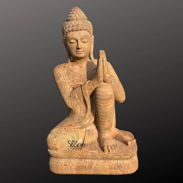 A majestic 6-foot Buddha sculpture, expertly carved from natural sandstone is prominently displayed against a plain dark gray background highlighting its intricate details and spiritual presence. The statue depicts Buddha in a kneeling posture with one knee raised and hands pressed together in the Anjali Mudra a gesture of reverence greeting, and prayer. His eyes are gently closed conveying a deep sense of peace humility and meditative calm. The sandstone material gives the sculpture a beautiful earthy reddish-brown hue with visible natural graining and a textured finish emphasizing its authentic stone origin and handcrafted artistry. Buddha's robes are finely sculpted with flowing undulating lines that suggest soft fabric gracefully draped over his body. His head is adorned with tightly curled hair an ushnisha at the crown signifying his spiritual wisdom and elongated earlobes. His facial features are rendered with exquisite serenity reflecting a profound inner tranquility. The sculpture rests on a multi-tiered carved base, also made of sandstone which features delicate traditional patterns along its edges providing a stable and aesthetically pleasing foundation. The posture itself conveys profound respect and devotion making this sandstone Buddha sculpture an ideal focal point for a sacred space meditation garden or any area designed to inspire peace and spiritual reflection. Its significant height and natural stone composition make it a durable and timeless piece of art embodying serenity and grace.