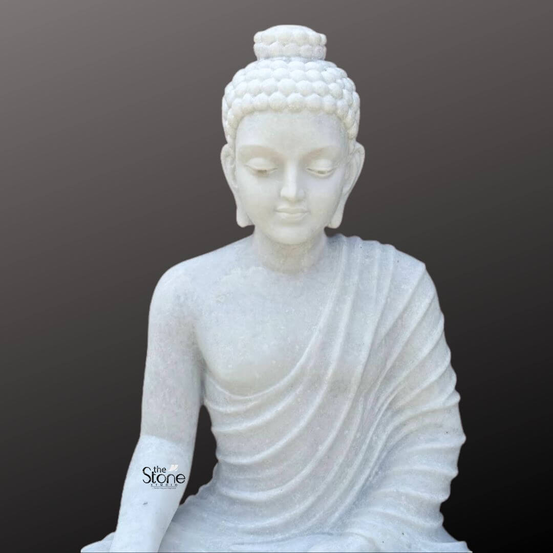 3 Feet Marble Buddha Idol 8 A beautifully detailed close-up of a premium three-foot marble Buddha sculpture is presented against a dark neutral background emphasizing its exquisite craftsmanship and serene expression. This image perfect for an article on a premium marble Buddha sculpture focuses on the upper body and face of the Buddha capturing the intricate artistry of the statue. The Buddha's head is slightly bowed and his eyes are gently closed or downcast conveying a profound state of inner peace meditation and spiritual tranquility. The sculpture showcases the luminous quality of the white marble which highlights the smooth contours of the Buddha's face and shoulders. His facial features are finely rendered with a calm and contemplative expression characterized by a slender nose well-defined lips, and elegantly arched eyebrows. A prominent ushnisha on his head symbolizing wisdom and spiritual attainment is meticulously carved with his hair sculpted into tight symmetrical curls. His elongated earlobes are clearly visible indicative of his princely past and renunciation of worldly possessions. The flowing robes are draped gracefully over his left shoulder and across his chest with naturalistic pleats that add depth and realism to the sculpture without detracting from its overall serene aesthetic. The details of the folds are expertly carved suggesting soft fabric while being rendered in hard marble. The smooth polished surface of the white marble softly reflects light, giving the statue a radiant quality that enhances its divine presence This close-up view effectively communicates the intricate details and the peaceful meditative essence of the premium three-foot marble Buddha sculpture making it a compelling image for any discussion of high-quality Buddhist art and spirituality.
