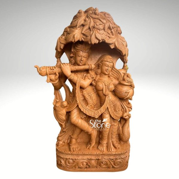 A beautifully sculpted 3.5-foot Radha Krishna statue carved from brown sandstone is presented against a clean softly diffused background that transitions from light grey to white making it an ideal choice for garden décor. The divine couple Radha and Krishna are depicted standing together under an intricately carved tree canopy with Krishna playing his flute. Lord Krishna stands gracefully on the left holding a flute to his lips captivating Radha with his divine music. He is adorned with an elaborate crown jewelry, and flowing garments, all meticulously carved with fine details. A peacock a symbol often associated with Krishna is depicted near his feet. Radha stands beside him on the right her expression one of devotion and enchantment holding a pot or pitcher symbolizing offerings or the abundance of nature. She is also adorned with a crown, necklaces, armlets, and a flowing skirt all intricately detailed in the brown sandstone. The most striking feature is the overarching tree canopy above them, with its dense realistic foliage carved in high relief providing a naturalistic frame and shelter for the divine couple. The entire 3.5-foot Radha Krishna statue rests on a sturdy multi-tiered rectangular base which is also adorned with traditional decorative motifs further enhancing its aesthetic appeal. The warm earthy tone and subtle texture of the brown sandstone give the sculpture a natural organic feel, perfectly blending with an outdoor garden environment. This detailed brown sandstone sculpture beautifully embodies love devotion, and harmony directly aligning with an article discussing Radha Krishna idols for garden décor.