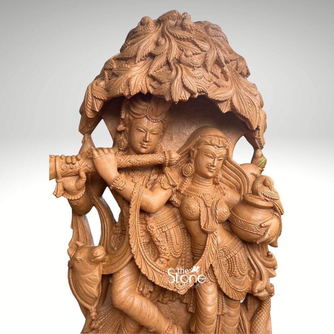 Radha Krishna Idol for Garden Décor 3.5ft 5 A magnificent 3.5-foot Radha Krishna Murti meticulously carved from rich brown sandstone with a distinctive tree design is presented against a clean softly diffused background that transitions from light grey to white making it an ideal centerpiece for a home temple. The divine couple Radha and Krishna are depicted standing gracefully together under an elaborate intricately sculpted tree canopy with Lord Krishna captivating Radha with his flute. Lord Krishna stands on the left, holding a flute to his lips his expression one of divine charm and musical enchantment. He is adorned with an ornate crown multiple necklaces armlets and flowing dhoti all rendered with exceptional detail in the brown sandstone. A sacred cow is intricately carved near his feet adding a pastoral and traditional element. Radha stands gracefully beside him on the right, her face expressing deep devotion and enchantment holding a pot or pitcher symbolizing offerings or the abundance of nature. She is also beautifully adorned with a crown necklaces and a flowing sari with all drapery and jewelry finely sculpted. The most striking feature is the dense overarching tree canopy above them meticulously carved with numerous leaves and branches in high relief from the same brown sandstone providing a naturalistic and sacred shelter. The entire 3.5-foot Radha Krishna Murti rests on a sturdy multi-tiered rectangular base which is richly decorated with traditional scrolling and floral motifs along its edges. The warm earthy tone and subtle texture of the brown sandstone give the sculpture a natural timeless and elegant aesthetic perfectly blending with various home temple designs. This detailed brown sandstone sculpture beautifully embodies eternal love devotion, and harmony directly aligning with an article discussing Radha Krishna Murti with a tree design for home temples.