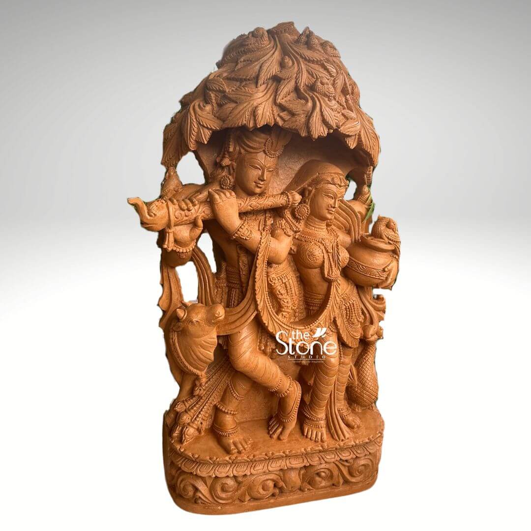Radha Krishna Idol for Garden Décor 3.5ft 4 A captivating 3.5-foot Radha Krishna sculpture meticulously carved from rich brown sandstone is prominently displayed against a clean softly diffused background that transitions from light grey to white. The divine couple Radha and Krishna are depicted standing gracefully together under an elaborate intricately sculpted tree canopy resembling a Kadamba tree with Lord Krishna enchanting Radha with his flute. Lord Krishna stands on the left, holding a flute to his lips his expression one of divine charm and musical enchantment. He is adorned with an ornate crown multiple necklaces armlets and flowing dhoti all rendered with exceptional detail in the brown sandstone. A sacred cow is intricately carved near his feet, adding a pastoral and traditional element to the scene. Radha stands gracefully beside him on the right, her face expressing deep devotion and enchantment holding a pot or pitcher symbolizing offerings or the abundance of nature. She is also beautifully adorned with a crown necklaces and a flowing sari with all drapery and jewelry finely sculpted. The most striking feature is the dense overarching tree canopy above them, meticulously carved with numerous leaves and branches in high relief from the same brown sandstone providing a naturalistic and sacred shelter. This detailed Kadamba tree design enhances the spiritual narrative. The entire 3.5-foot Radha Krishna sculpture rests on a sturdy, multi-tiered rectangular base which is richly decorated with traditional scrolling and floral motifs along its edges. The warm, earthy tone and subtle texture of the brown sandstone give the sculpture a natural, timeless, and elegant aesthetic. This detailed brown sandstone sculpture beautifully embodies eternal love devotion, and harmony, directly aligning with an article discussing Radha Krishna sculptures under a Kadamba tree.