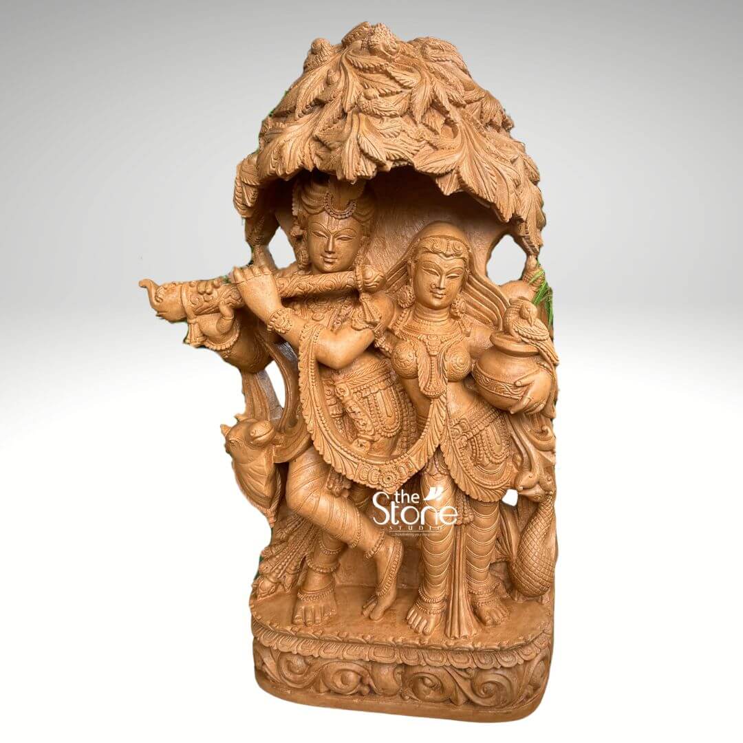 Radha Krishna Idol for Garden Décor 3.5ft 7 A charming 3.5-foot Radha Krishna statue, intricately carved from brown sandstone with an integrated tree design is prominently displayed against a clean softly diffused background that transitions from light grey to white making it a perfect addition to a garden. The divine couple Radha and Krishna are depicted standing gracefully together under an elaborate meticulously sculpted tree canopy with Lord Krishna enchanting Radha with his flute. Lord Krishna stands on the left, holding a flute to his lips his expression one of divine charm and musical enchantment. He is adorned with an ornate crown multiple necklaces armlets and flowing dhoti all rendered with exceptional detail in the brown sandstone. A sacred cow is intricately carved near his feet, adding a pastoral and traditional element to the scene. Radha stands gracefully beside him on the right her face expressing deep devotion and enchantment holding a pot or pitcher symbolizing offerings or the abundance of nature. She is also beautifully adorned with a crown necklaces and a flowing sari with all drapery and jewelry finely sculpted. The most striking feature is the dense overarching tree canopy above them meticulously carved with numerous leaves and branches in high relief from the same brown sandstone providing a naturalistic and sacred shelter. The entire 3.5-foot Radha Krishna statue rests on a sturdy multi-tiered rectangular base which is richly decorated with traditional scrolling and floral motifs along its edges. The warm earthy tone and subtle texture of the brown sandstone give the sculpture a natural timeless, and elegant aesthetic perfectly blending with various garden landscapes. This detailed brown sandstone sculpture beautifully embodies eternal love devotion, and harmony directly aligning with an article discussing Radha Krishna statues with a tree design suitable for gardens.