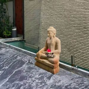 A serene two-foot tall Buddha statue meticulously carved from off-white sandstone sits in a meditative posture holding a delicate pink lotus flower in its hands. The statue is positioned outdoors on a textured brown base next to a modern water feature with small green tiles. The Buddha's calm expression tightly coiled hair and flowing robes are rendered with exquisite detail highlighting the natural grain and subtle variations in the sandstone. The statue is set against a textured wall with horizontal grooves creating a contemporary backdrop that contrasts with the traditional form of the Buddha. In the foreground a stylish black and white veined marble floor extends adding to the modern aesthetic of the space. Small subtle outdoor lights are visible on the marble floor suggesting illumination during evening hours. Lush green plants in the background add a touch of natural vitality to the scene. This image beautifully captures the harmonious blend of spiritual tranquility and modern design showcasing how a stone Buddha statue can enhance the peaceful ambiance of a contemporary outdoor living area or garden in Hyderabad. The presence of the lotus flower further emphasizes purity love, and spiritual awakening making this statue not just a decorative piece but a profound symbol of peace and enlightenment.