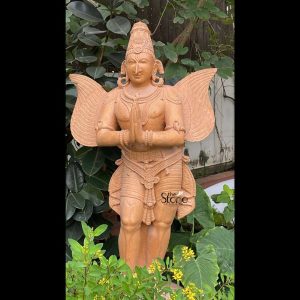 A magnificent 5-foot Stone Garuda statue meticulously carved from sandstone stands prominently amidst lush greenery in a garden setting highlighting its cultural significance possibly in Ahmedabad. The statue depicts Garuda the mythical divine eagle or bird-like creature vahana of Lord Vishnu, in a humanoid form with powerful wings. He is shown standing reverently with his hands pressed together in the Anjali Mudra a gesture of prayer respect and devotion. The sandstone material gives the sculpture a beautiful natural earthy reddish-brown hue with subtle textures and visible natural graining emphasizing its authentic stone origin and handcrafted artistry. Garuda is richly adorned with elaborate finely carved details representing traditional jewelry and attire. These include a magnificent tiered crown with intricate patterns ornate necklaces around his neck armlets on his upper arms and bracelets on his wrists. A sacred thread drapes across his bare torso and a highly decorative dhoti or lower garment is intricately sculpted with patterns. The most striking features are his large powerful wings spread out behind him meticulously detailed with feather-like textures symbolizing his ability to soar through the heavens. His face features expressive eyes a prominent nose and a serene, devoted expression. Elongated earlobes with traditional earrings are also visible. The statue stands on an integrated base partially obscured by the foreground plants. It is surrounded by various green leafy plants and small yellow flowering bushes creating a natural and vibrant backdrop. This sandstone Garuda statue with its significant height and intricate details serves as an inspiring and powerful symbol of devotion protection and divine strength perfect for a garden or temple entrance.