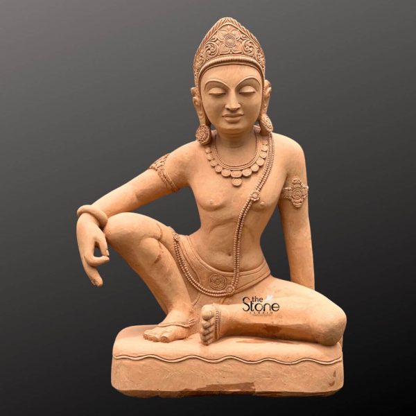 A majestic 6-foot Stone Indra statue meticulously carved from sandstone is prominently displayed against a plain dark gray background highlighting its unique posture and divine regalia. The statue depicts Lord Indra the king of Svarga and the Devas in a relaxed squatting or half-seated posture with his left knee raised and right leg extended or bent underneath. His left arm rests casually on his raised knee while his right arm extends forward. His eyes are gently closed conveying a sense of serene meditation wisdom, and profound calm. The sandstone material gives the sculpture a beautiful natural earthy reddish-brown hue with subtle textures and visible natural graining emphasizing its authentic stone origin and handcrafted artistry. Indra is richly adorned with elaborate finely carved details representing traditional jewelry and attire. These include a magnificent multi-tiered crown with intricate patterns ornate necklaces around his neck armlets on his upper arms and bracelets on his wrists and ankles. A sacred thread drapes across his torso. His muscular yet graceful physique is evident with flowing lines suggesting his divine form. His facial features are rendered with exquisite serenity reflecting a powerful yet peaceful presence. The entire figure rests on a simple sturdy sandstone base which provides a stable and aesthetically pleasing foundation. This unique posture and the rich ornamentation make this sandstone Indra statue an ideal focal point for a large garden, temple or any space seeking to embody divine power, wisdom and tranquility through traditional Indian sculpture. Its significant height and natural stone composition make it a durable and enduring piece of art.