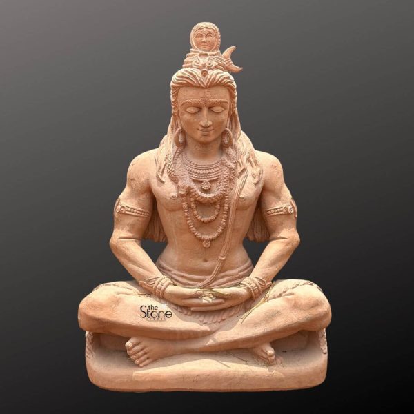 A majestic 2.5-foot tall Lord Shiva statue meticulously carved from reddish-brown sandstone sits in a profound meditative posture ideal for a garden setting. Lord Shiva is depicted in deep contemplation with his eyes gently closed exuding an aura of divine serenity and inner peace. His body is strong and well-defined adorned with traditional elaborate jewelry including multiple necklaces armlets and a sacred thread across his torso. His long matted hair flows gracefully over his shoulders and at the top of his head a small figure of Ganga is visible within his topknot symbolizing the descent of the Ganges through his hair. His hands are placed in his lap in the Dhyana Mudra conveying ultimate tranquility. The natural texture and warm earthy tones of the sandstone lend an ancient and organic quality to the sculpture with subtle variations in color that enhance its natural beauty and depth making it blend seamlessly with natural surroundings. The statue rests on a simple integrated base that elevates it slightly. Presented against a plain dark grey background the warm hue and intricate details of the sandstone Shiva statue are beautifully highlighted. Its impressive 2.5-foot height makes it a significant focal point perfect for a meditation garden or a spiritual corner in any outdoor space inviting a sense of calm, devotion and divine presence. This Shiva statue serves as both a beautiful artistic element and a powerful symbol of meditation creation and destruction.